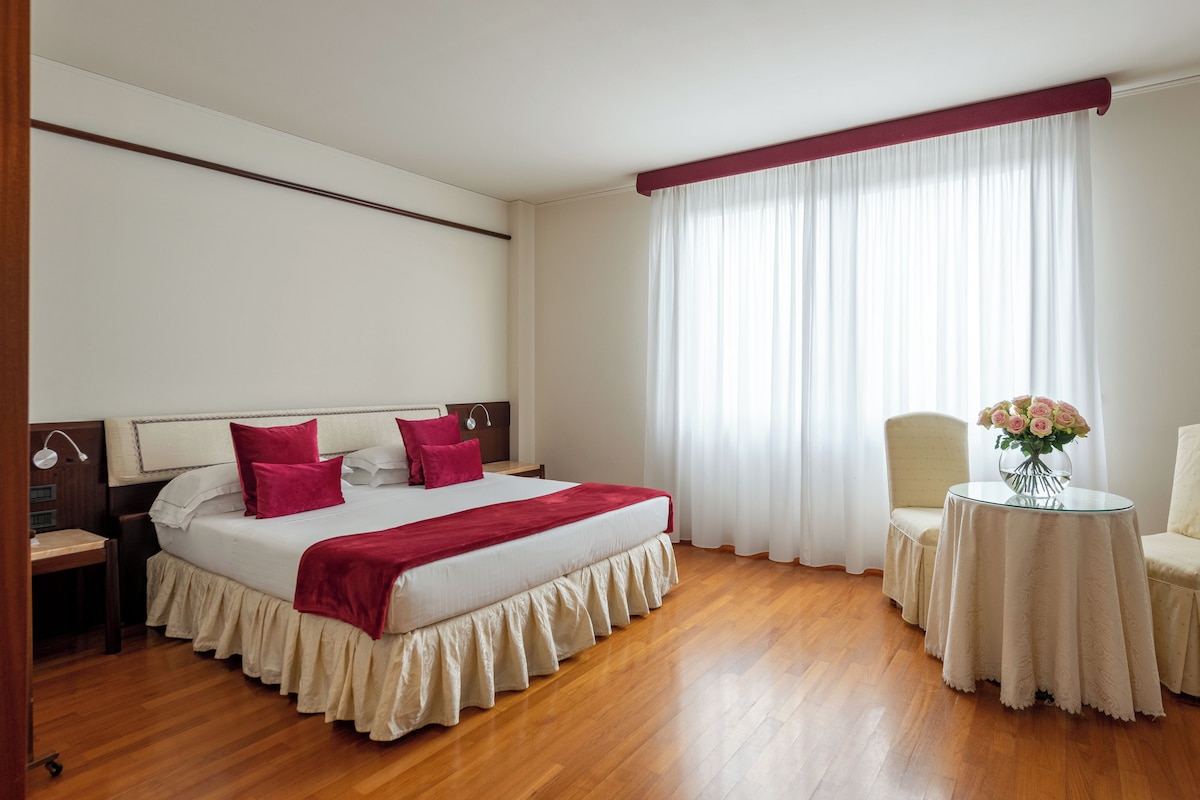 Habitación de hotel con una cama grande, un escritorio, una silla y una mesita de noche con un jarrón de flores.