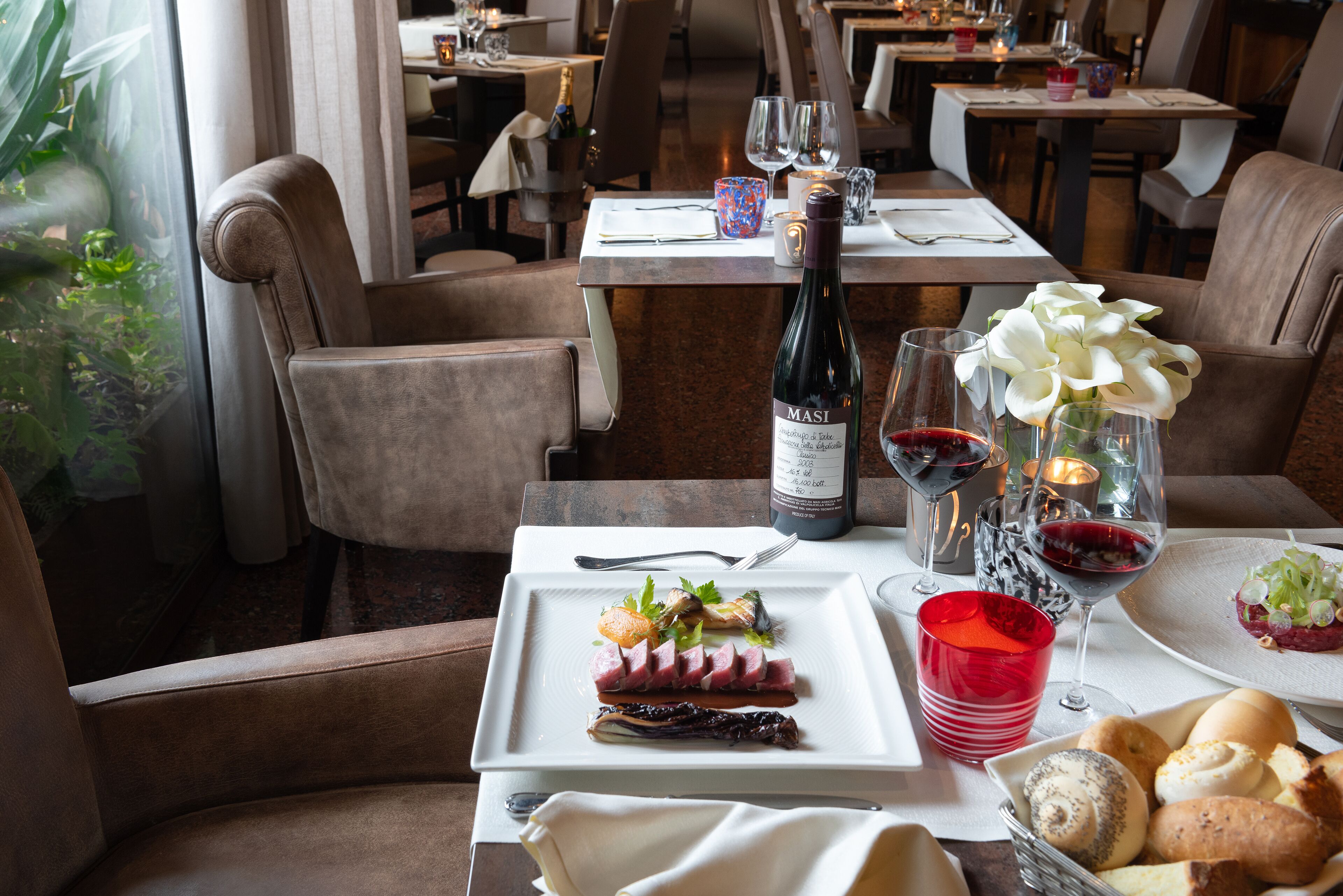 A dining table set with a bottle of Masi wine, red wine glasses, a red cup, a plate of food, and a basket of bread.