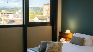 Ett hotellrum med en säng, kuddar, ett sängbord med en lampa och utsikt över byggnader genom ett fönster.