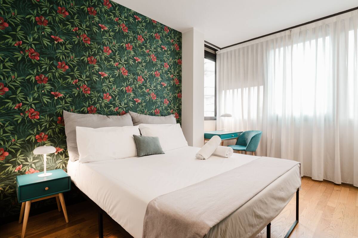 A neatly made bed with white linens, a teal bedside table, and a nightstand with a lamp. A window with sheer curtains allows natural light into the room.