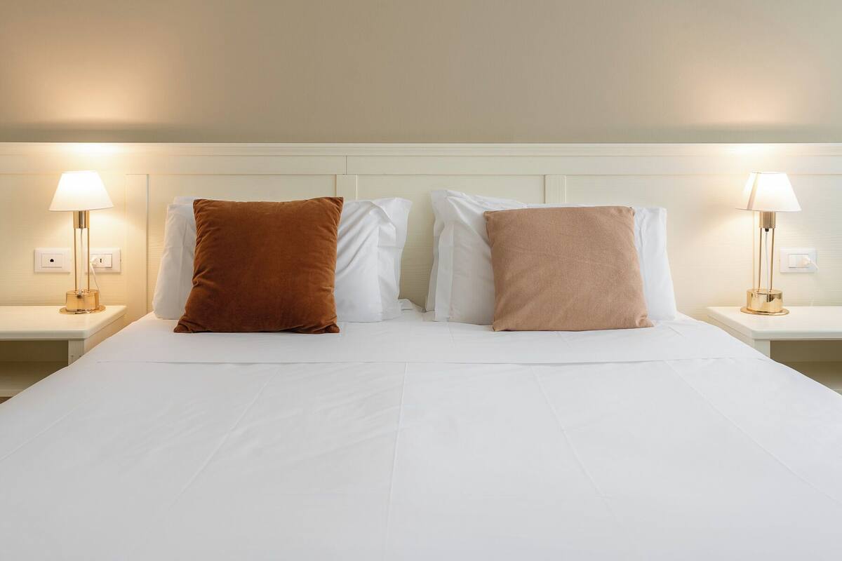 A neatly made bed with white bedding and two brown pillows, flanked by two bedside lamps on white nightstands.
