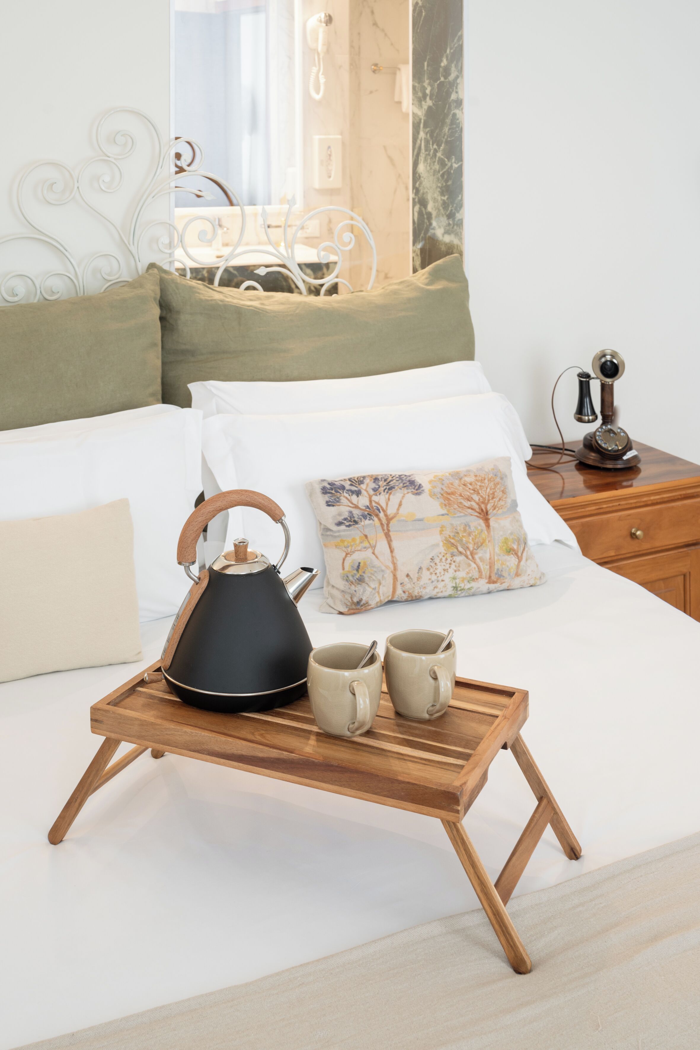 Un lit bien fait, sur lequel se trouve un plateau avec une bouilloire et deux tasses, un coussin décoratif et une table de chevet avec un téléphone.