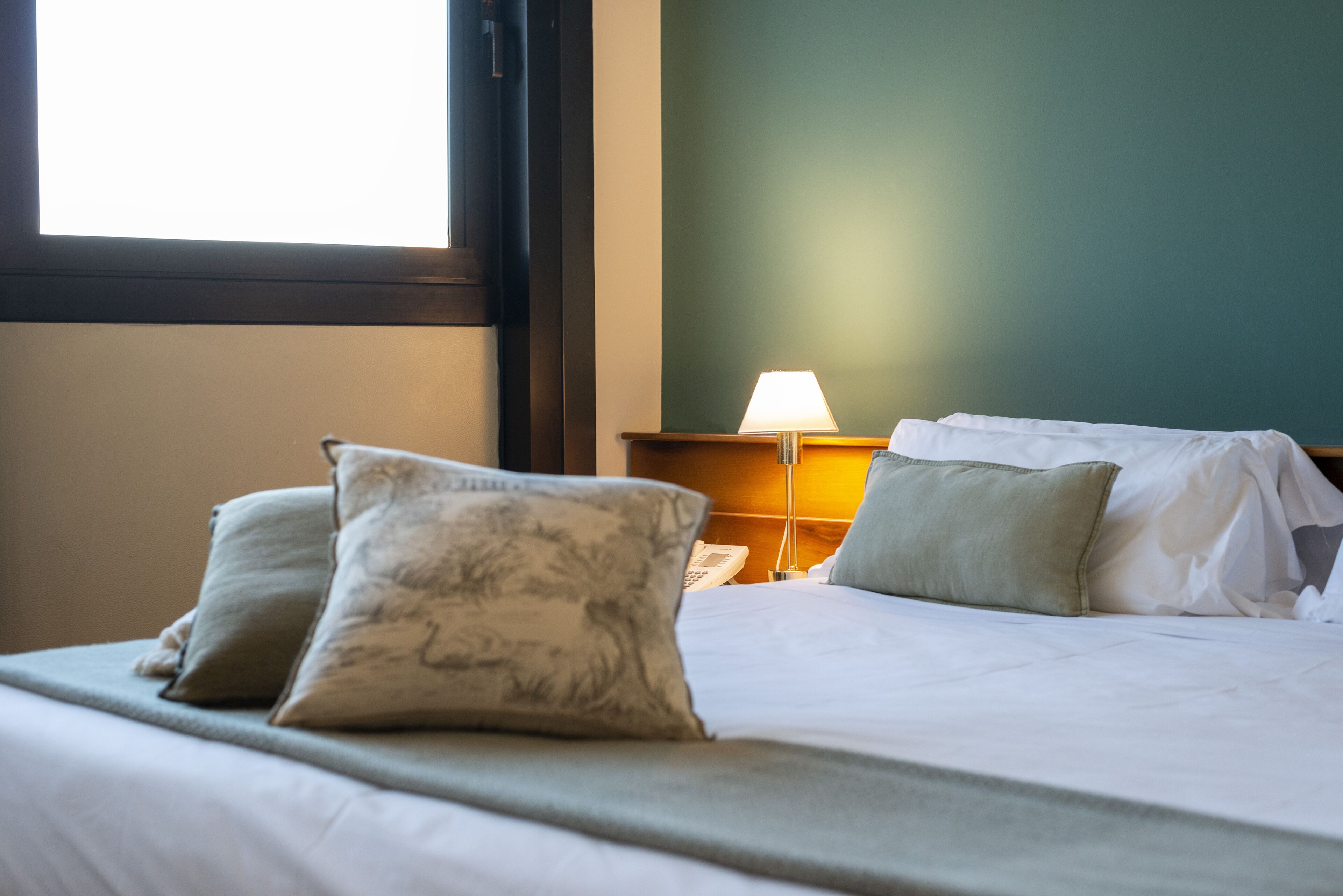 Quarto de hotel com cama, almofadas, abajur e mesa de cabeceira.