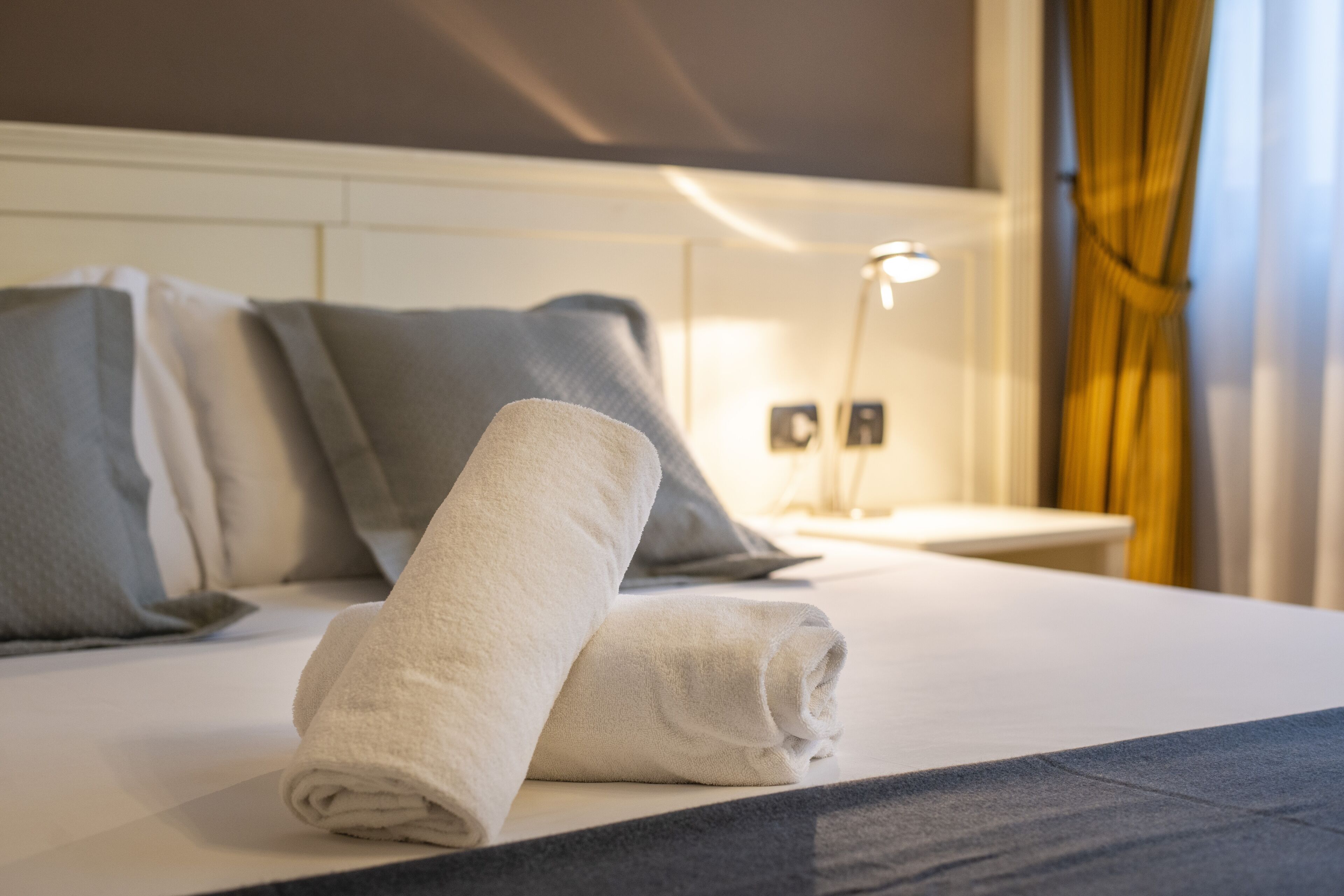 A neatly made bed with a rolled white towel, grey pillows, and a bedside table with a lamp.