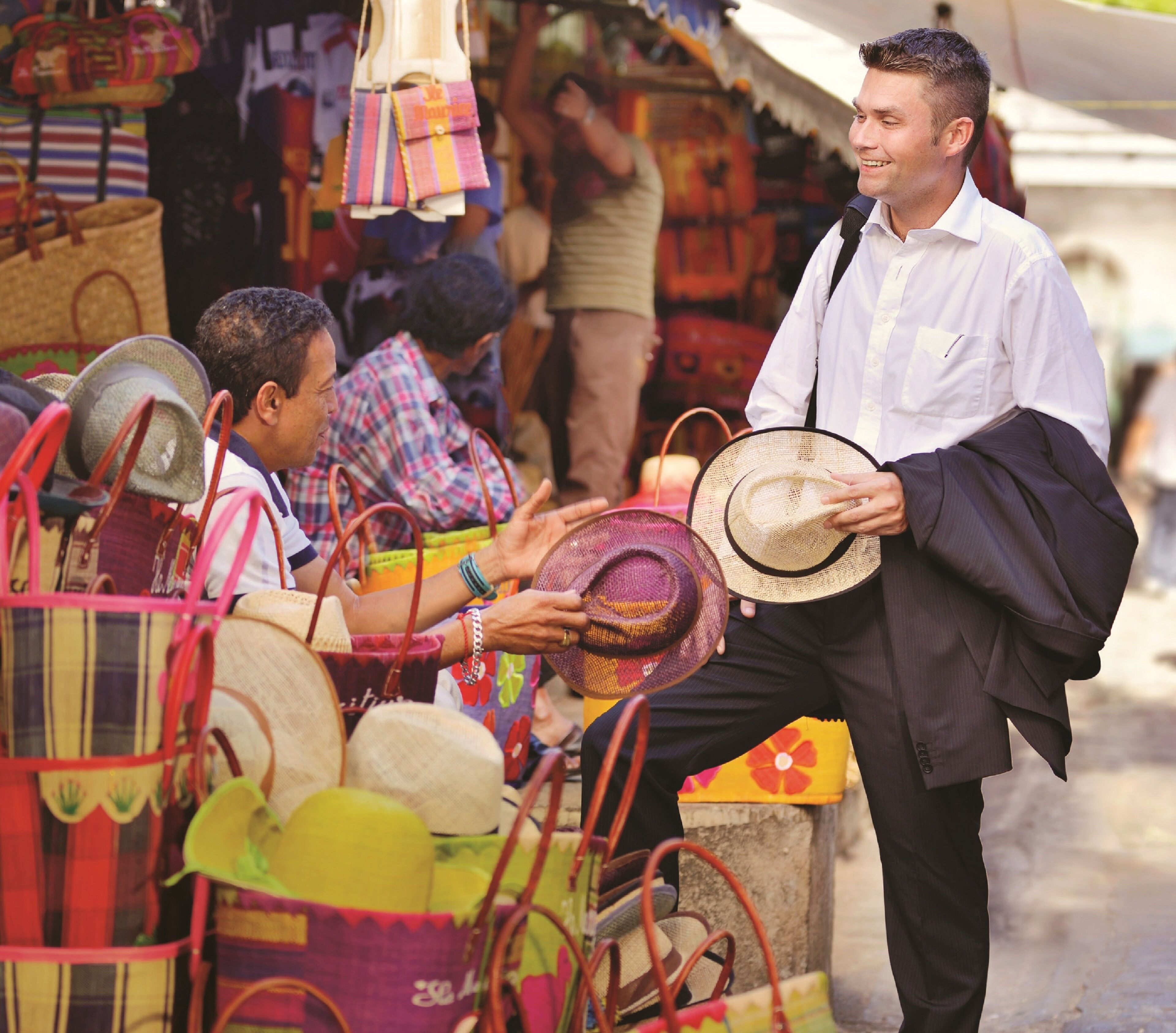 A man holding a hat and a bag, standing in a market with colorful bags and hats displayed.
