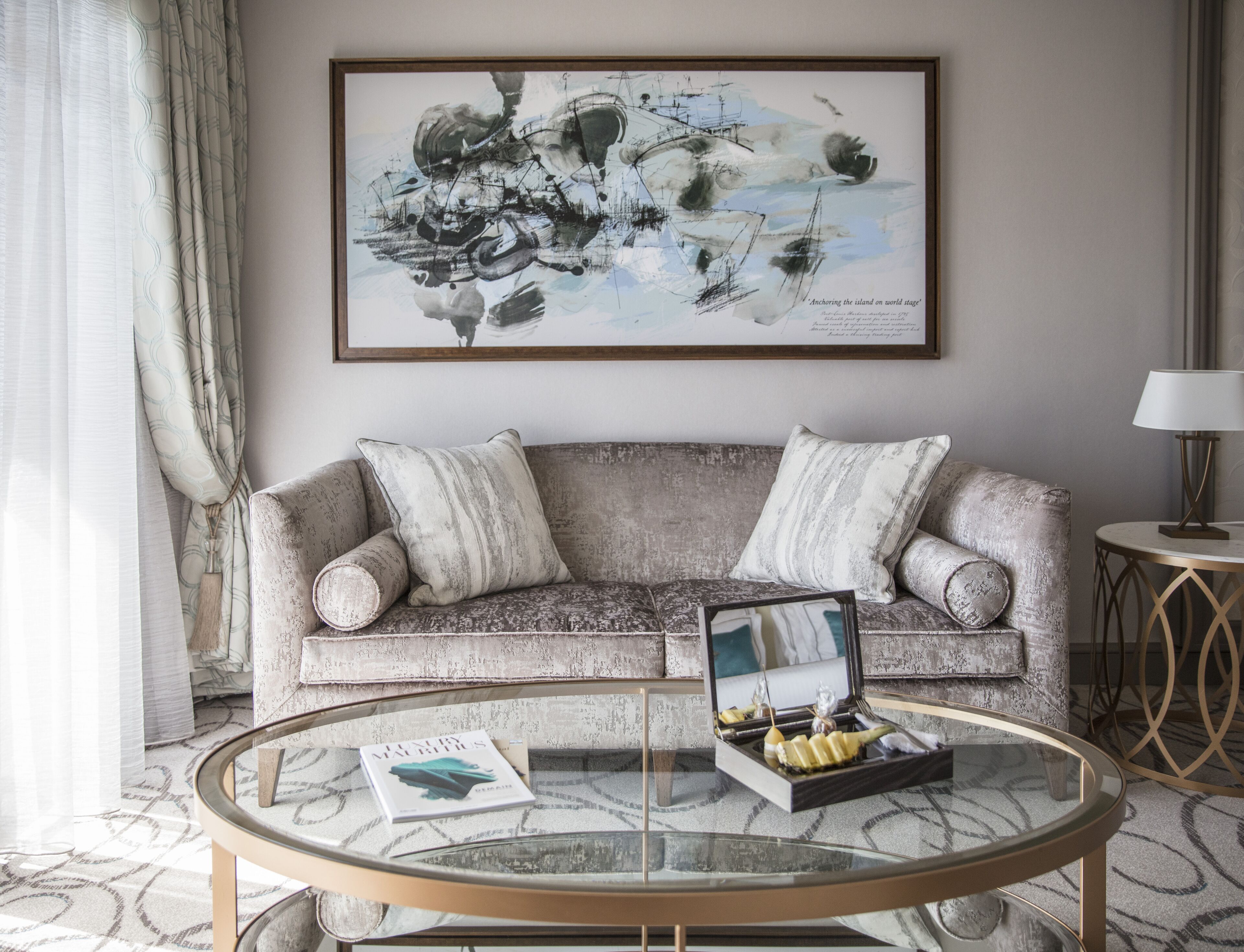 Una sala de estar con un sofá gris, dos cojines, una mesa de centro de cristal con un libro y objetos decorativos, y un cuadro abstracto enmarcado en la pared.