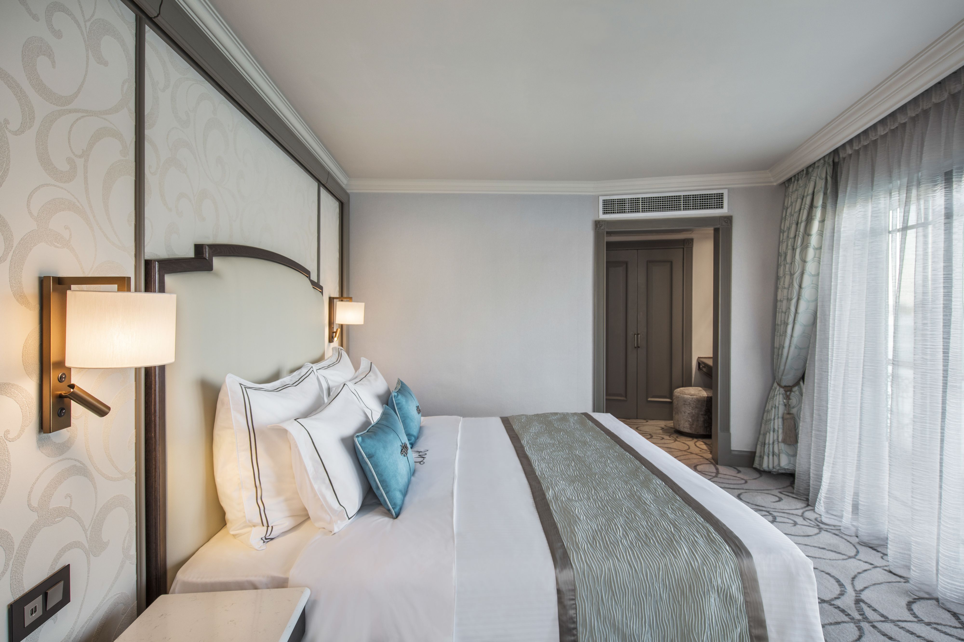 Une chambre d’hôtel avec un grand lit, deux lampes de chevet, une tête de lit dotée d’un luminaire, une petite table et une porte donnant sur une salle de bain.