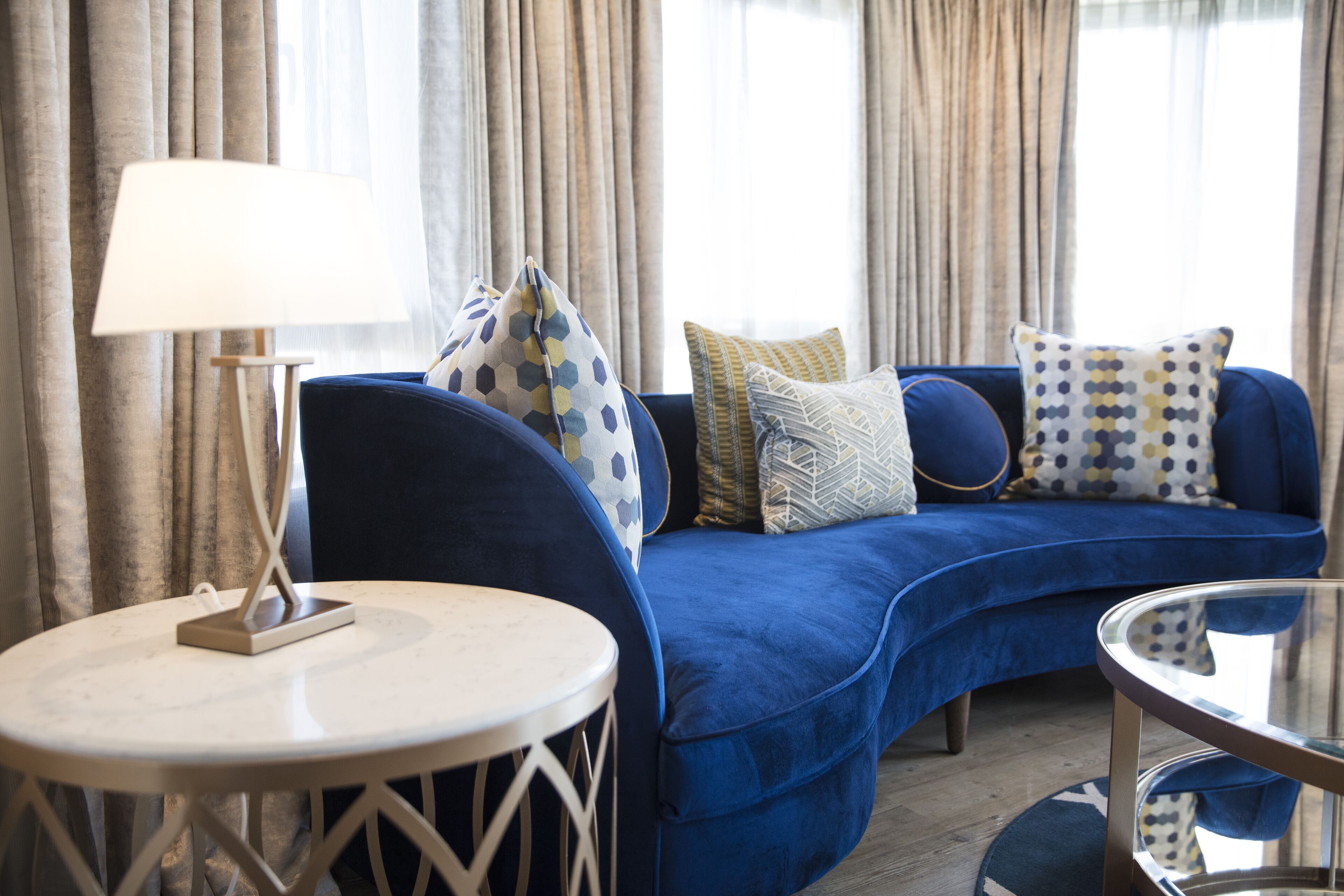 A blue sofa with patterned cushions, a round white table, and a lamp in a room with curtains and a window.