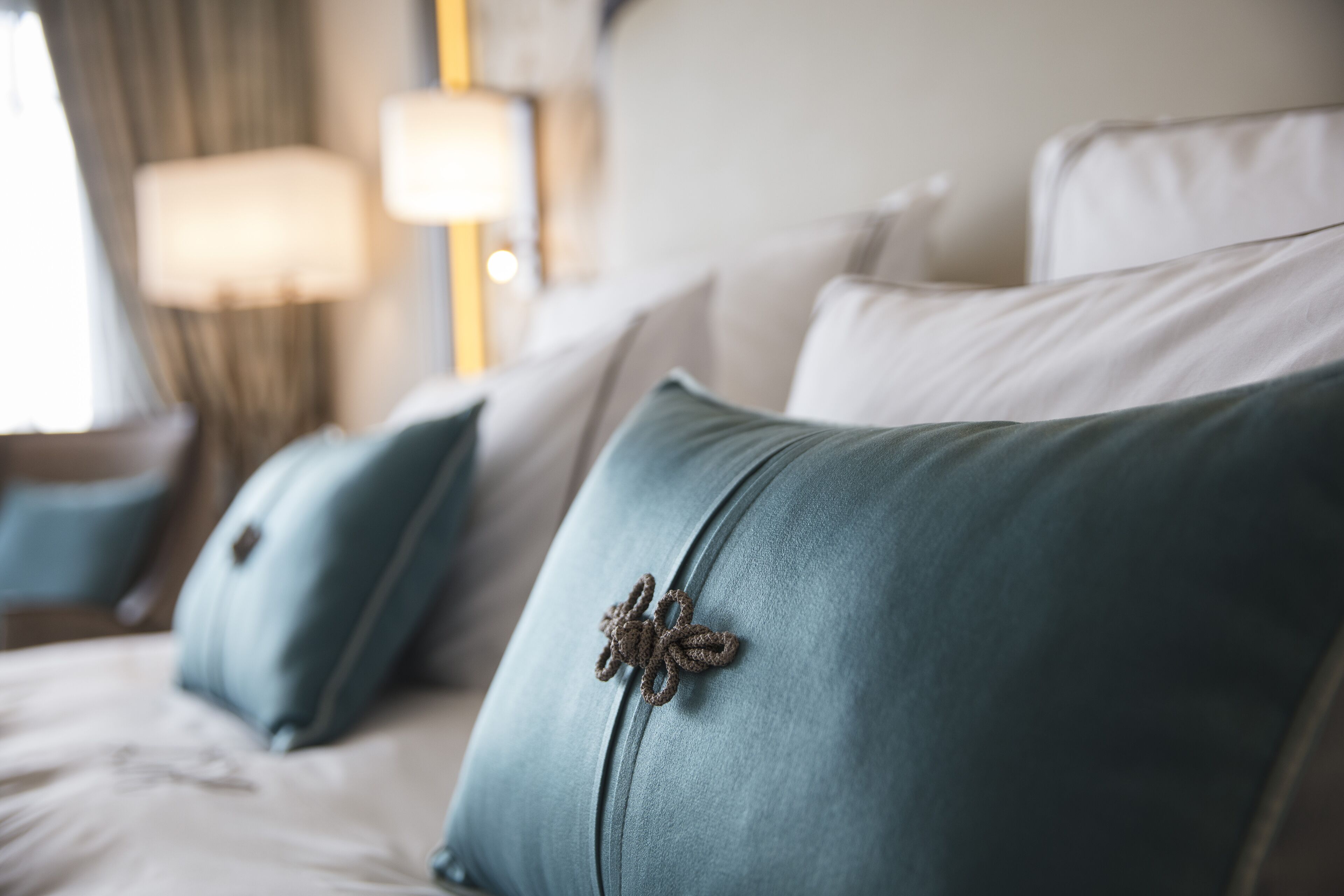 Un lit avec des coussins de couleur turquoise et une broche décorative, une lampe et une fenêtre avec des rideaux.