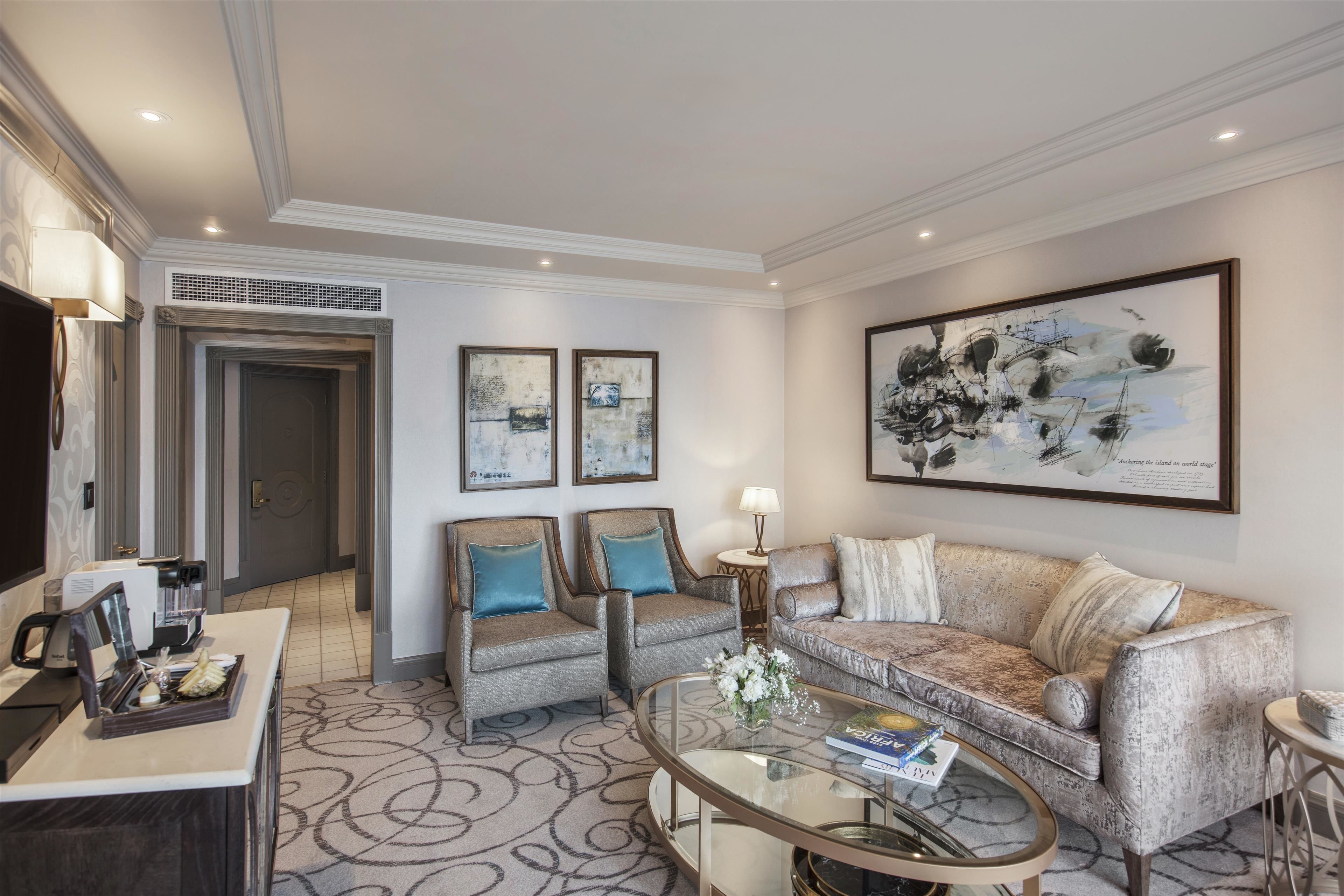 A modern living room with a sofa, two armchairs, a coffee table, and a TV. There are framed artworks on the walls and a coffee machine on the counter.