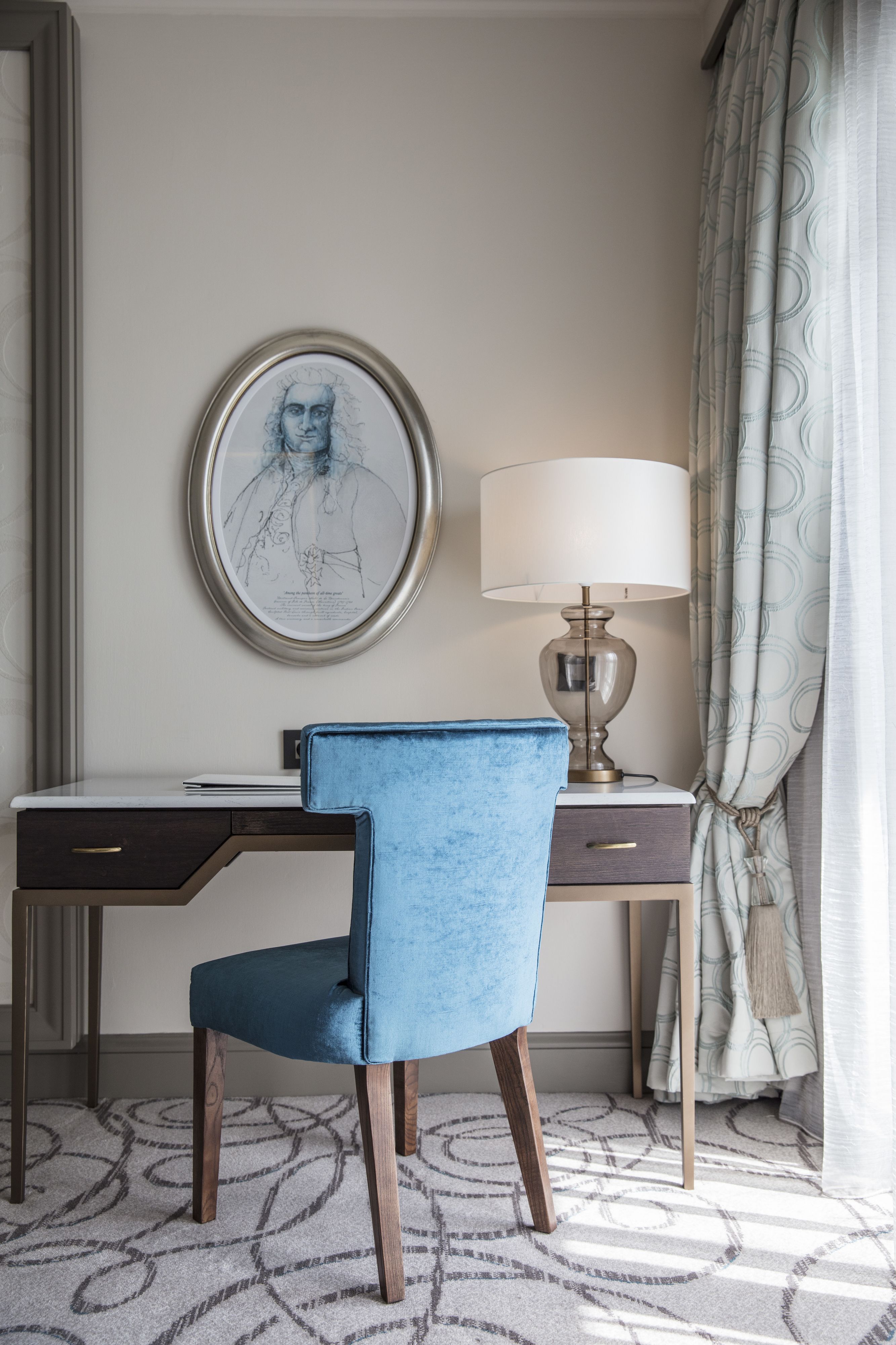 Una habitación con un escritorio, una silla, una lámpara, una obra de arte enmarcada y una alfombra estampada.