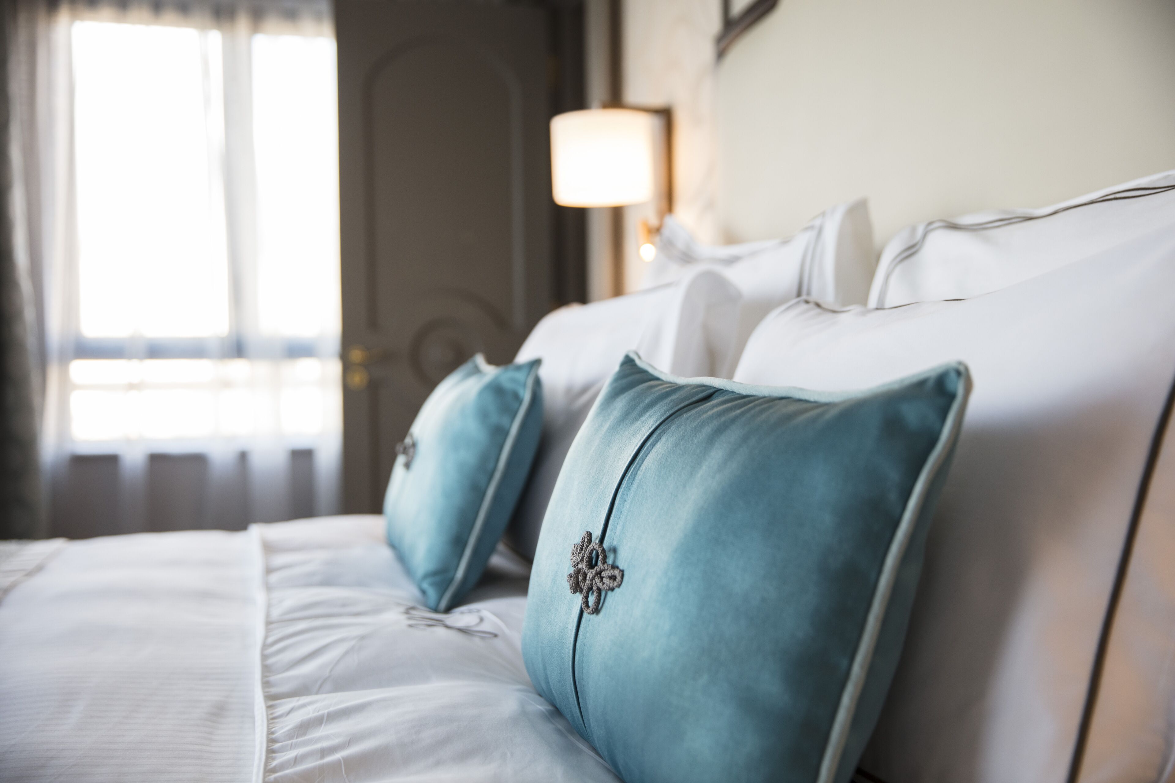 A neatly made bed with white linens and blue pillows, a bedside lamp, and a window with curtains.
