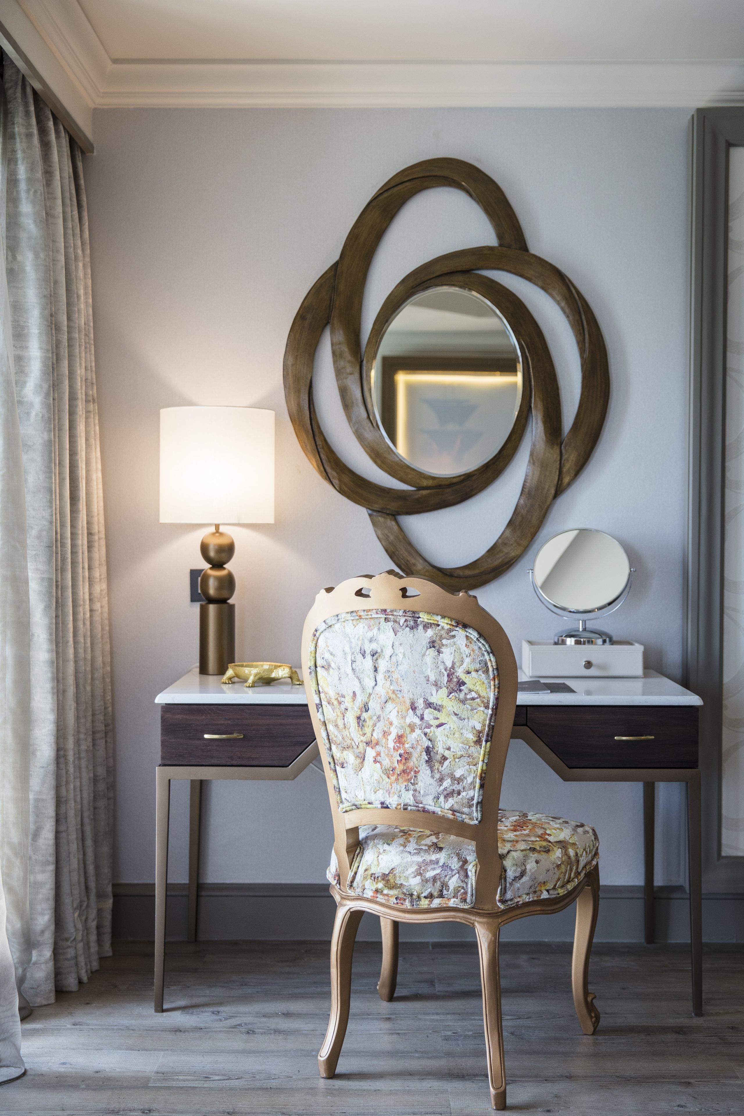 Una habitación con una silla de madera tapizada con motivos florales, un escritorio con una lámpara y un espejo montado en la pared.