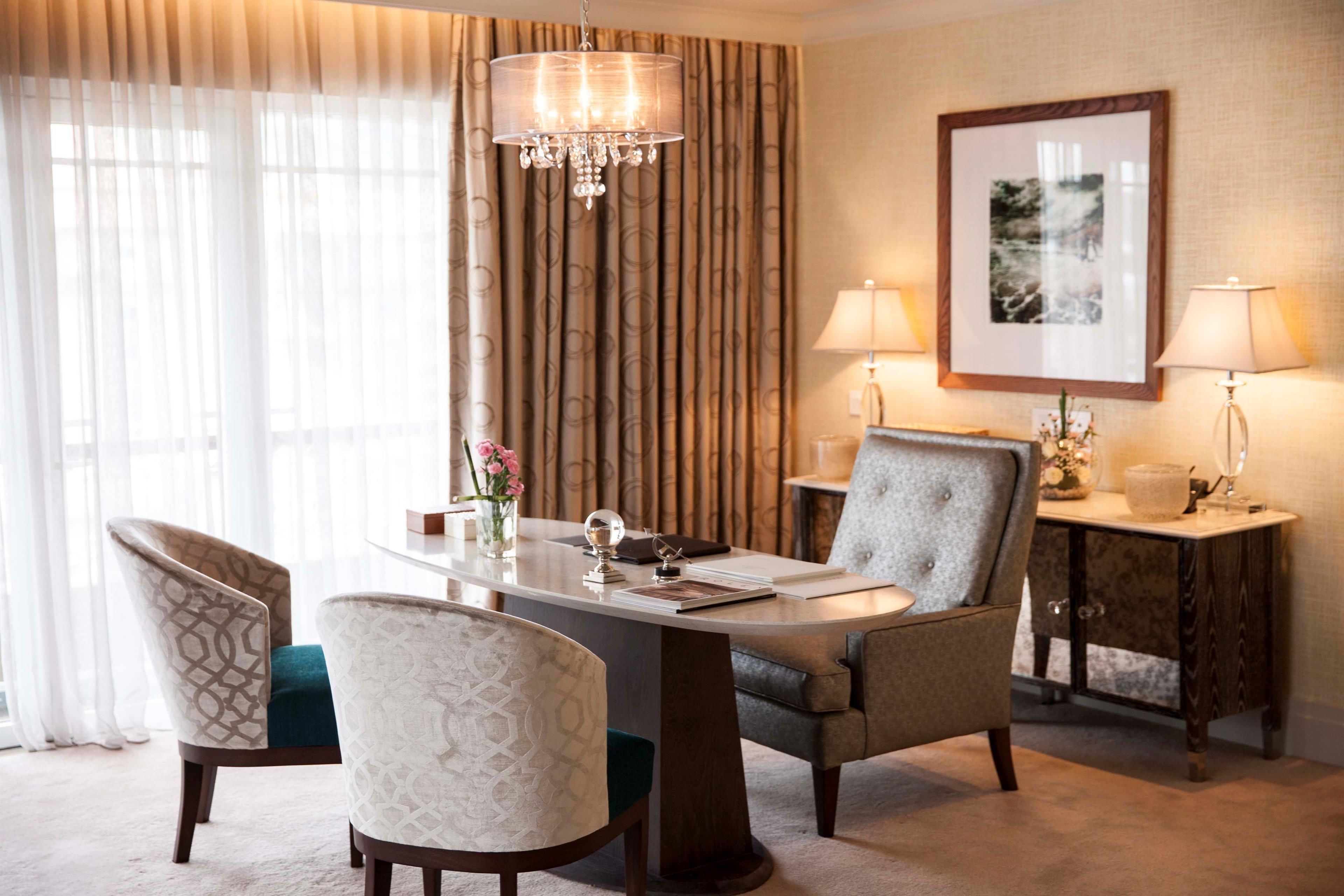 Une chambre d’hôtel comprenant une table à manger, deux chaises, un lustre, une table d’appoint avec une lampe et un tableau encadré au mur.