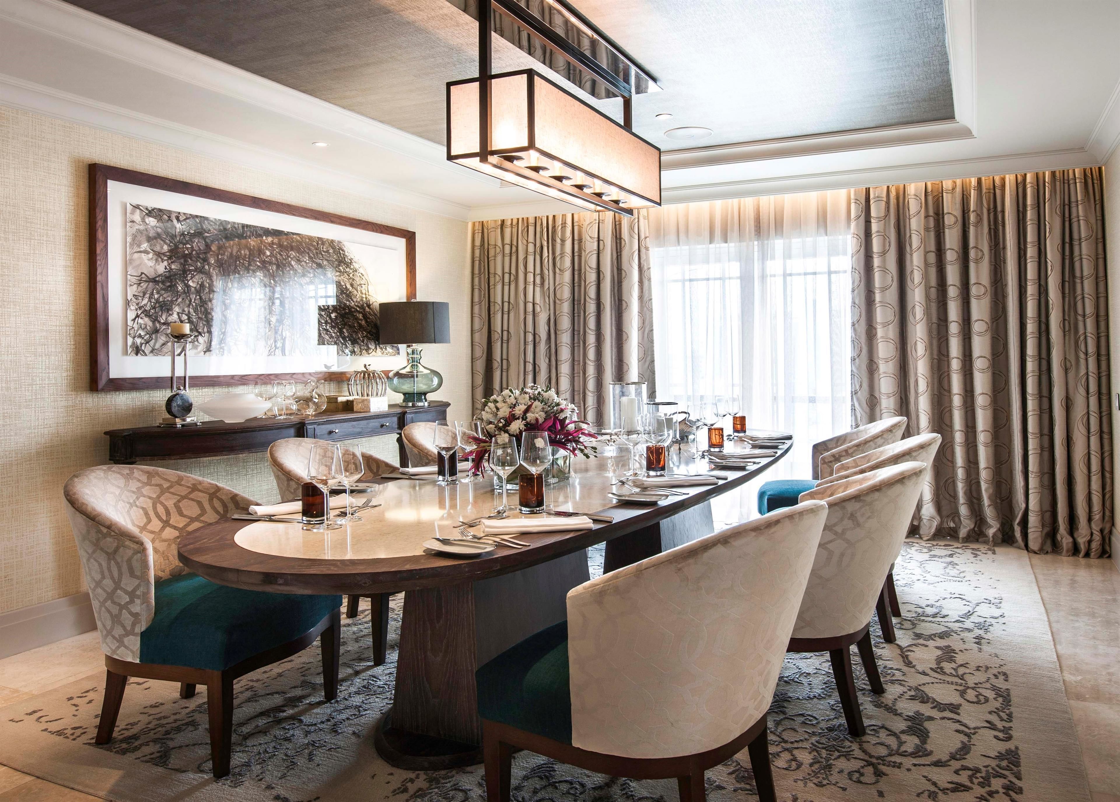 Uma sala de jantar com uma mesa comprida posta para seis pessoas, rodeada por cadeiras estofadas. Um candelabro pendurado acima e um quadro na parede.