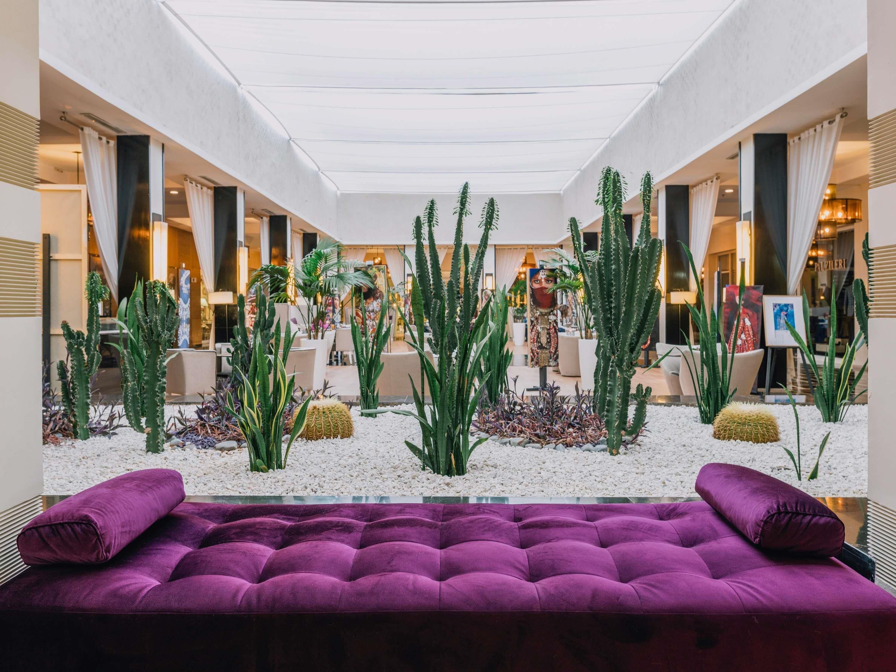 Un hall d’hôtel moderne avec un espace salon central agrémenté d’un canapé violet, entouré d’une variété de cactus et de plantes, et surmonté d’un plafond en verre.