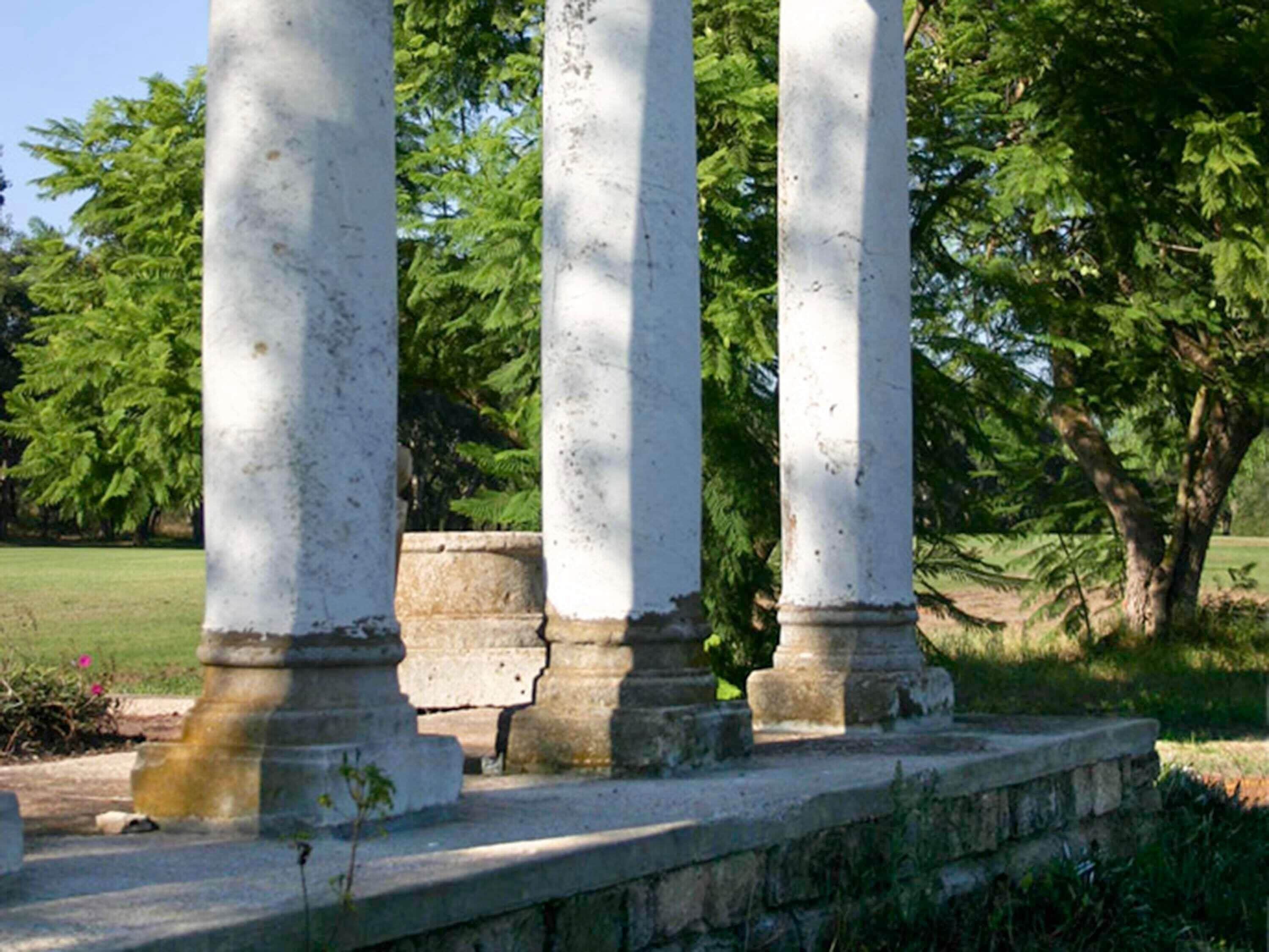 A set of white columns with stone bases, set in a grassy area with trees in the background.