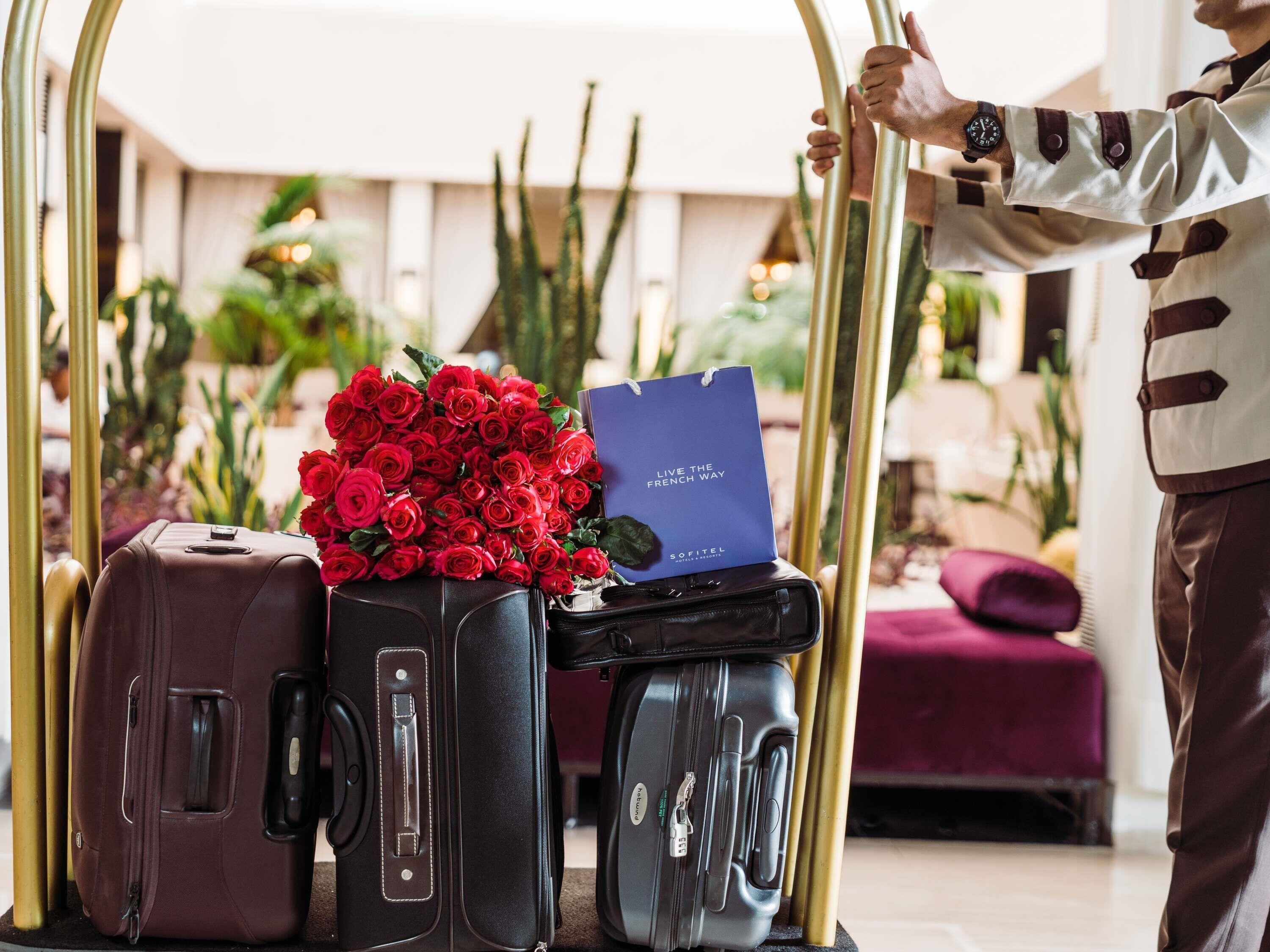 Une personne portant un t-shirt rayé, debout à côté d’un chariot à bagages, tenant un bouquet de roses rouges et une carte bleue.