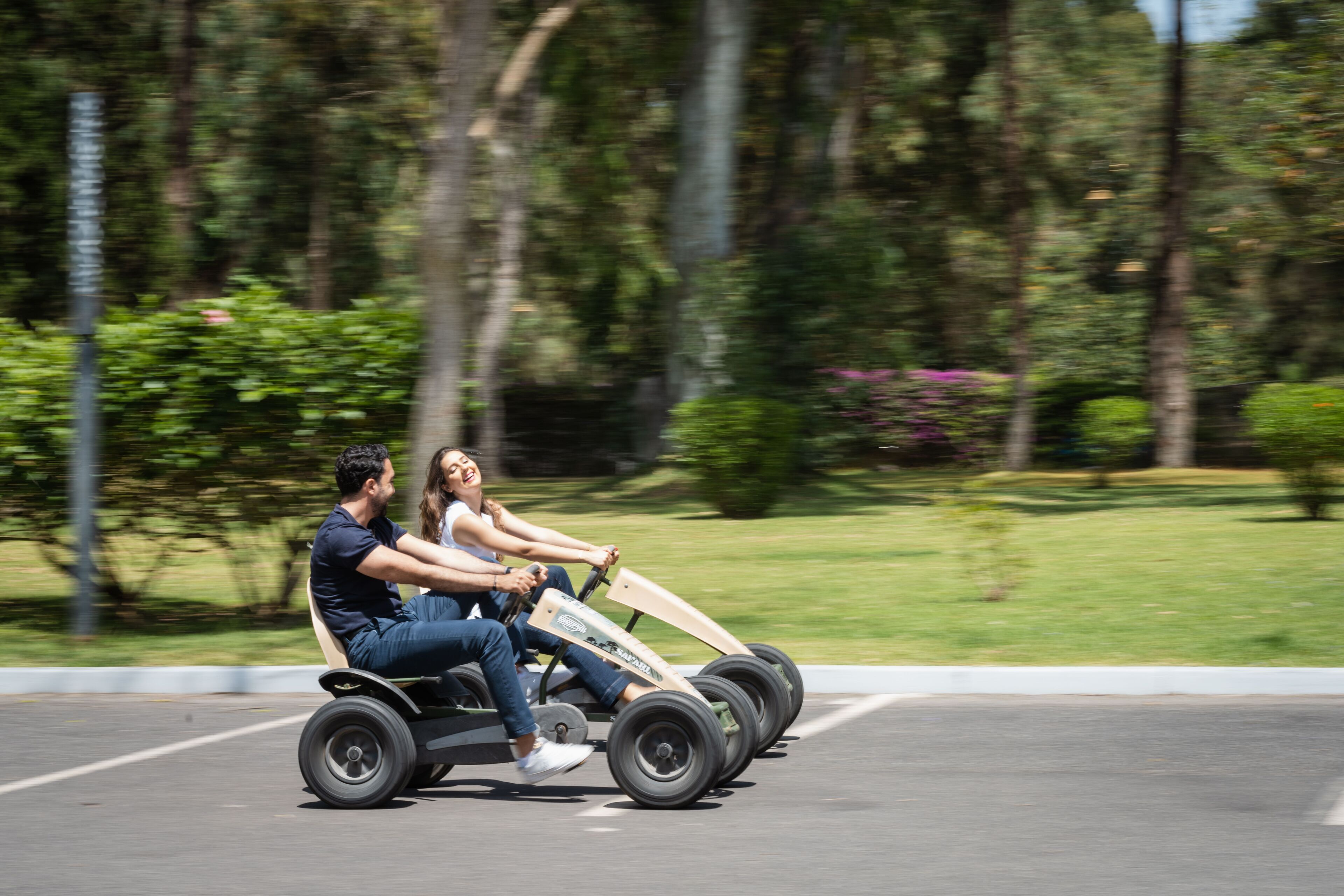 Dos personas en un karting en un estacionamiento, con árboles y césped al fondo.