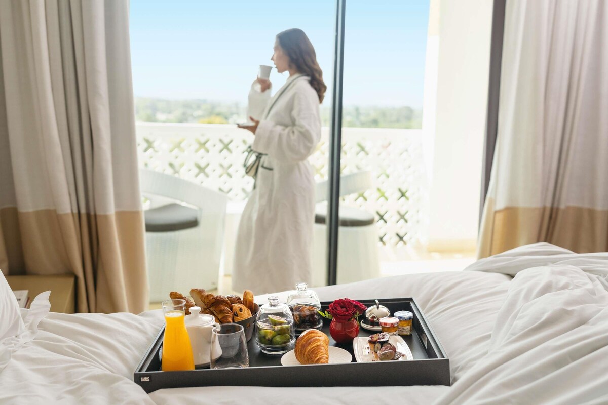 Ett frukostbricka med croissanter, bakverk, sylt och juice, placerat på sängen i ett hotellrum med utsikt från balkongen.