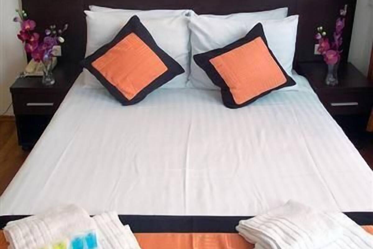 A neatly made bed with white linens and two orange pillows, a bedside table with a vase of flowers, and a wall-mounted light fixture.