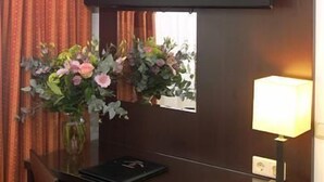 A hotel room with a TV mounted on the wall, a desk with a chair, a vase of flowers, and a lamp. - Hotel Falcon Plaza (Amsterdam)