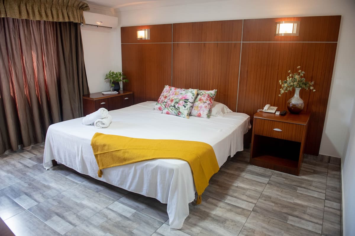 Quarto de hotel com uma cama grande, cabeceira de madeira, mesa de cabeceira com telefone e vaso, e janela com cortinas.