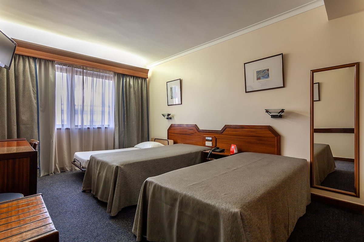 A hotel room with two single beds, a wooden desk, a chair, a bedside table with a lamp, a mirror, and a window with curtains.