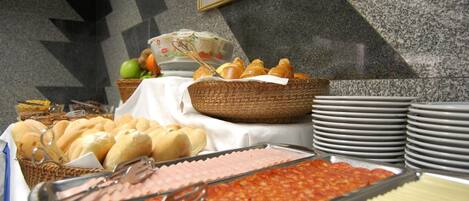 Un buffet proposant une variété de plats, notamment du pain, des pâtisseries et un plat de viande, disposés sur une table avec des assiettes empilées et un panier.