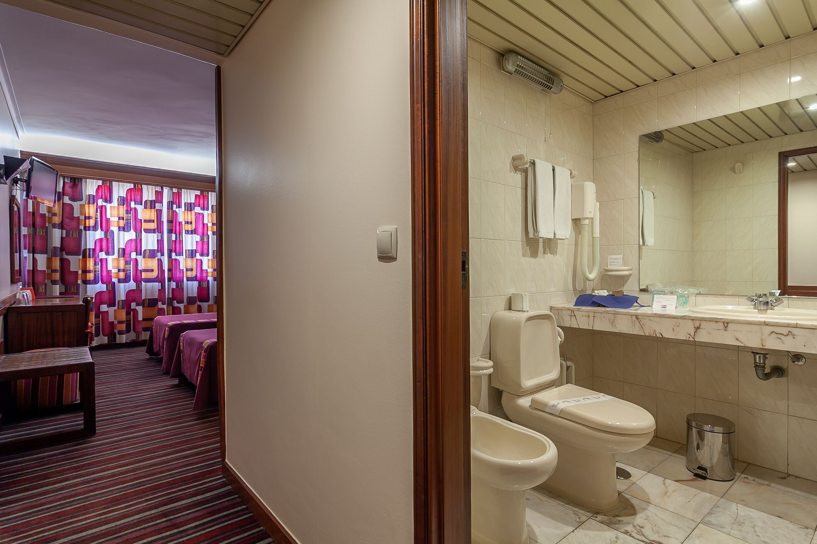 A hotel bathroom with a toilet, bidet, and sink. There is a trash can and a mirror above the sink.