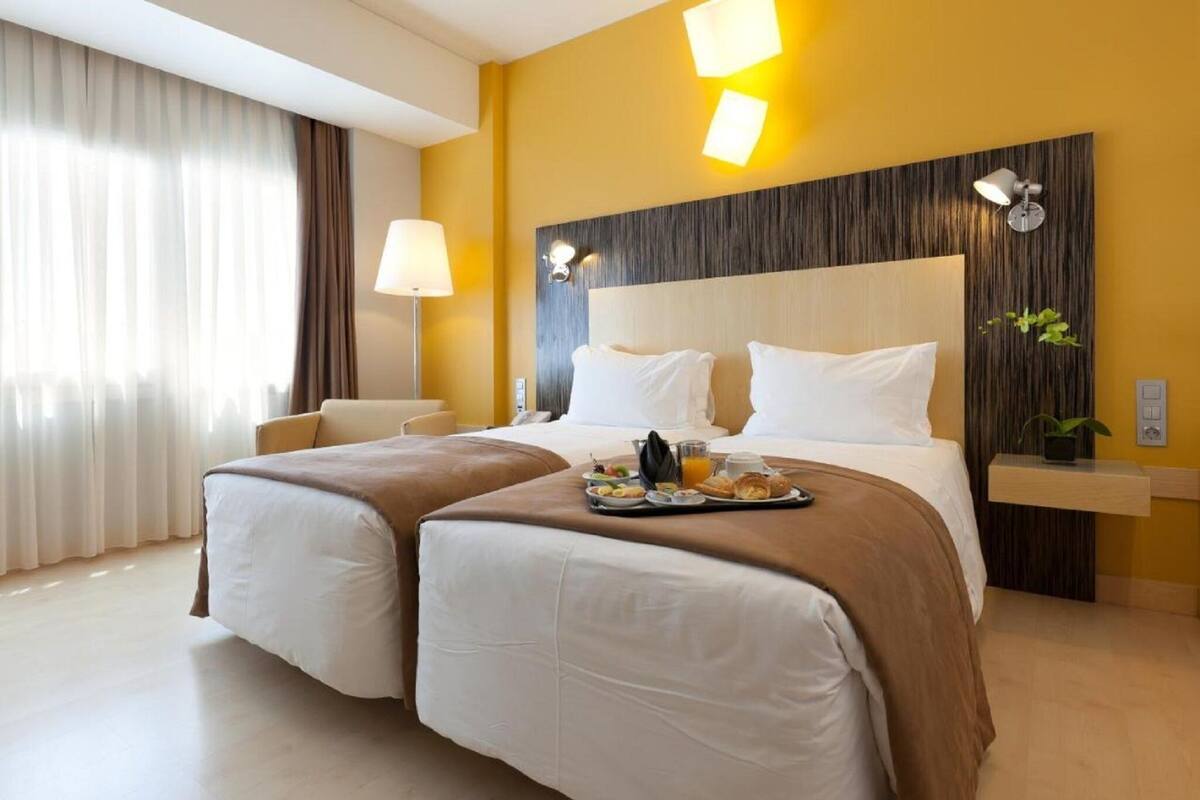 A hotel room with two beds, a bedside table, a lamp, and a tray with breakfast items.