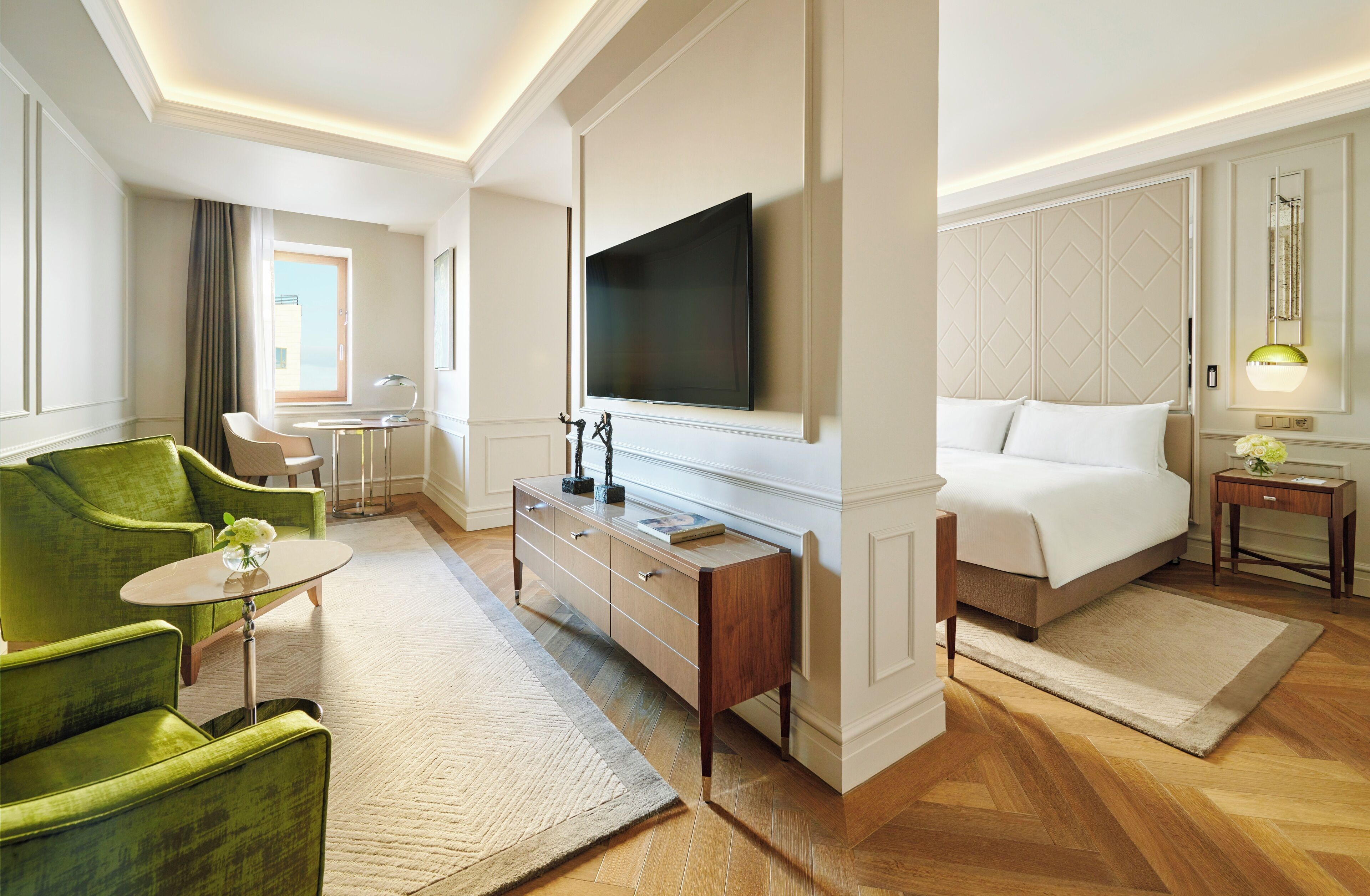 Quarto de hotel com uma cama grande, televisão na parede, uma poltrona verde, secretária com luminária e janela com cortinas.