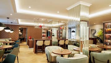 Un moderno vestíbulo de hotel con varias zonas de estar, incluyendo mesas redondas y sofás, y una partición de vidrio decorativa en el centro.