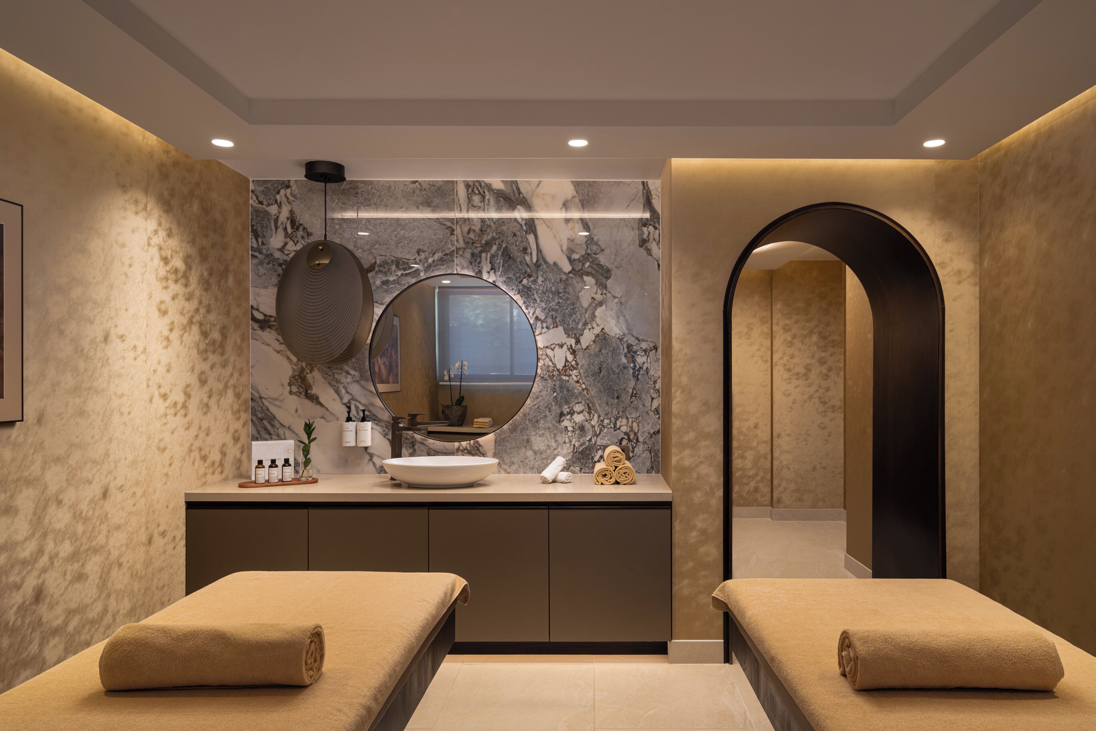 A modern spa room with two massage tables, a marble wall, a round mirror, and a sink with toiletries.