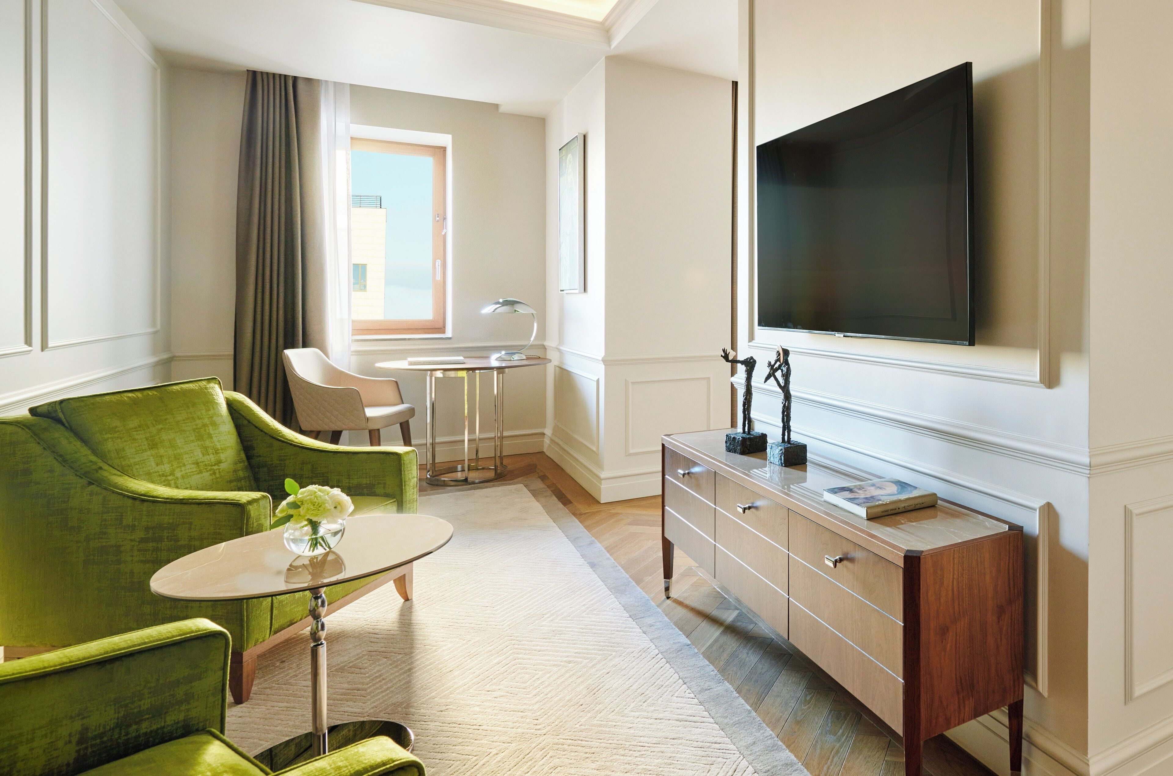 A hotel room with a green armchair, a small round table with a vase, a wooden dresser, a flat-screen TV, a desk with a lamp, and a window with curtains.