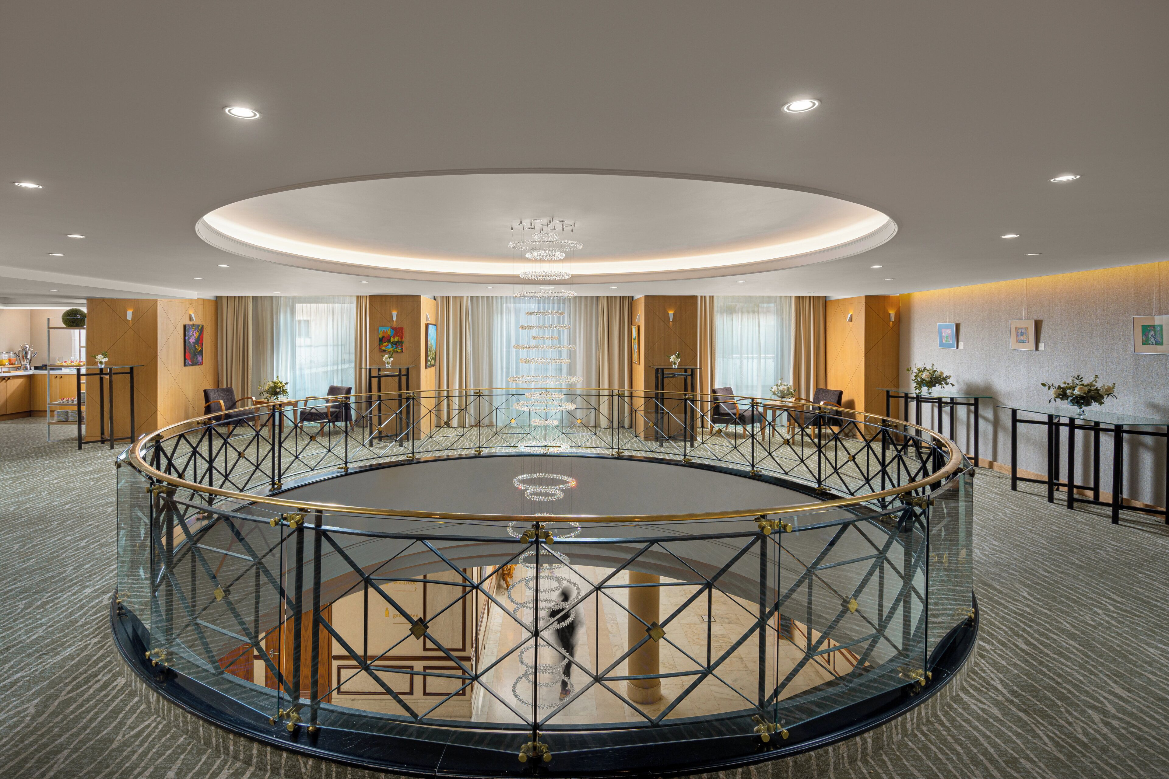 Um espaço amplo e circular com piso de vidro, cercado por uma grade de metal, e um lustre central.