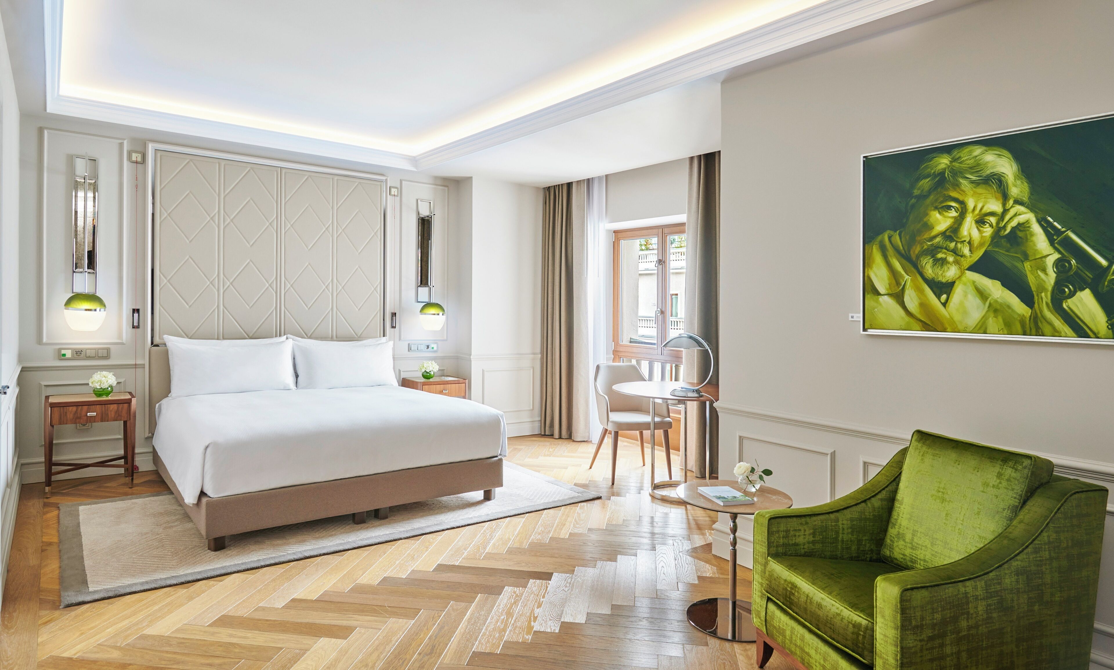 A modern hotel room with a large bed, a green armchair, a small table, and a painting on the wall.