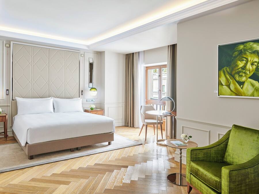 A modern hotel room with a large bed, a green armchair, a small table, and a painting on the wall.