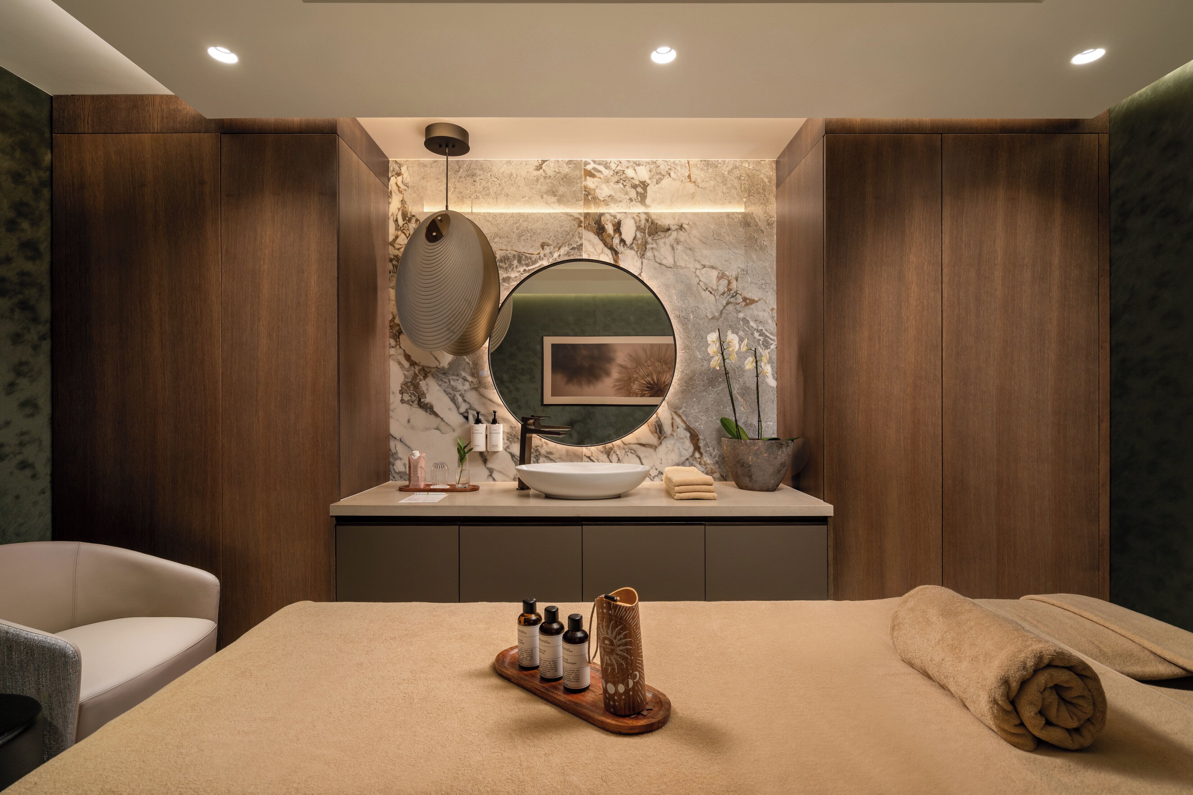 Un bagno moderno con un mobile vanitario in legno, uno specchio rotondo, uno schienale in marmo e un letto con asciugamano e articoli da toilette.