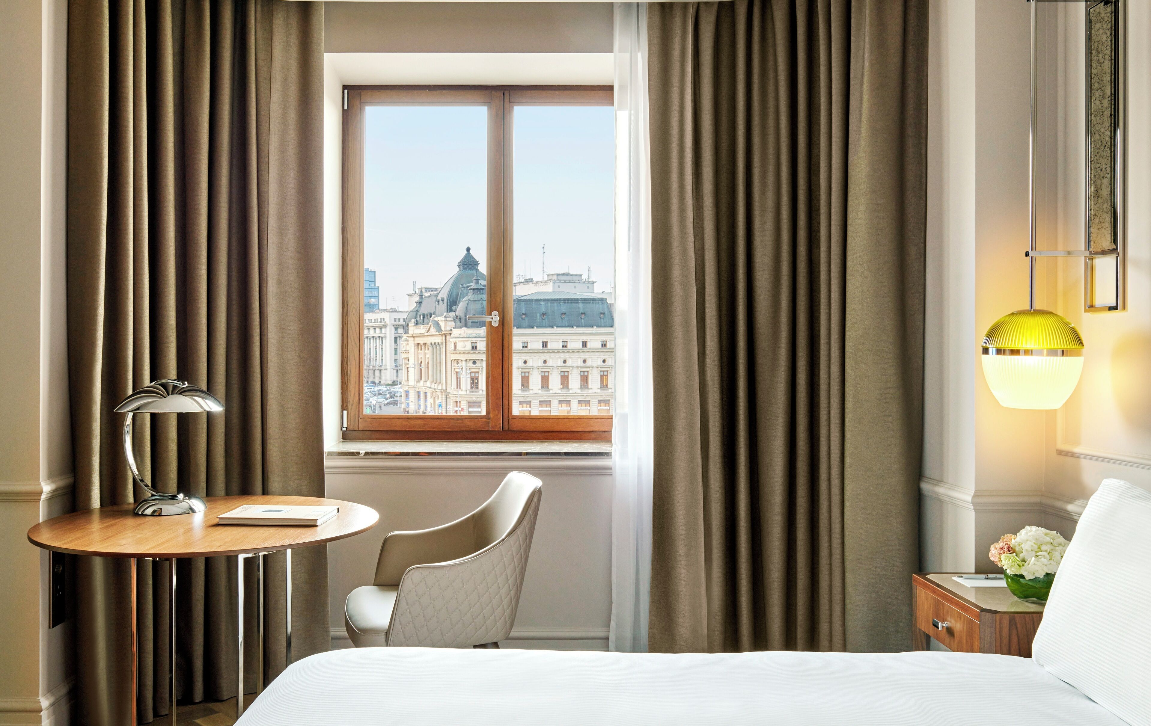 Habitación de hotel con cama, escritorio con lámpara, silla, ventana con cortinas y vista a la ciudad.