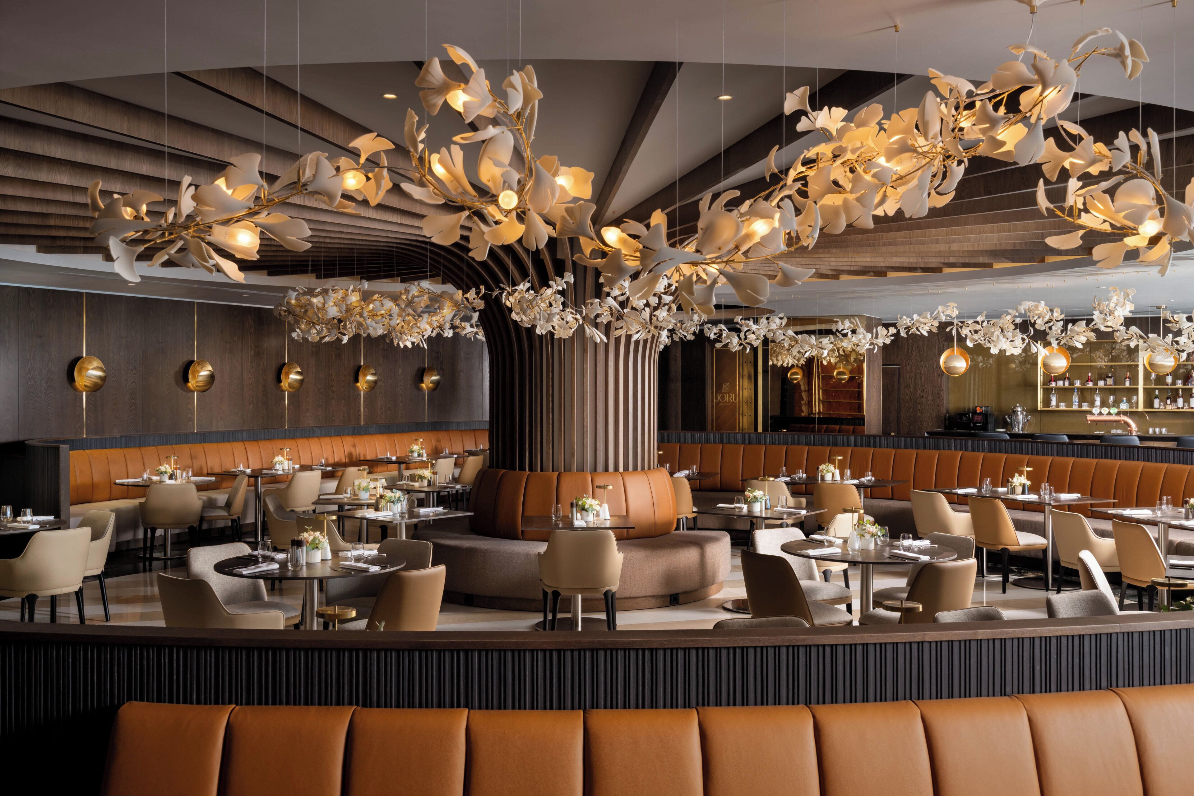 Ein modernes Restaurant mit einer zentralen Sitzgruppe, mehreren Esstischen und einem auffälligen Kronleuchter an der Decke.