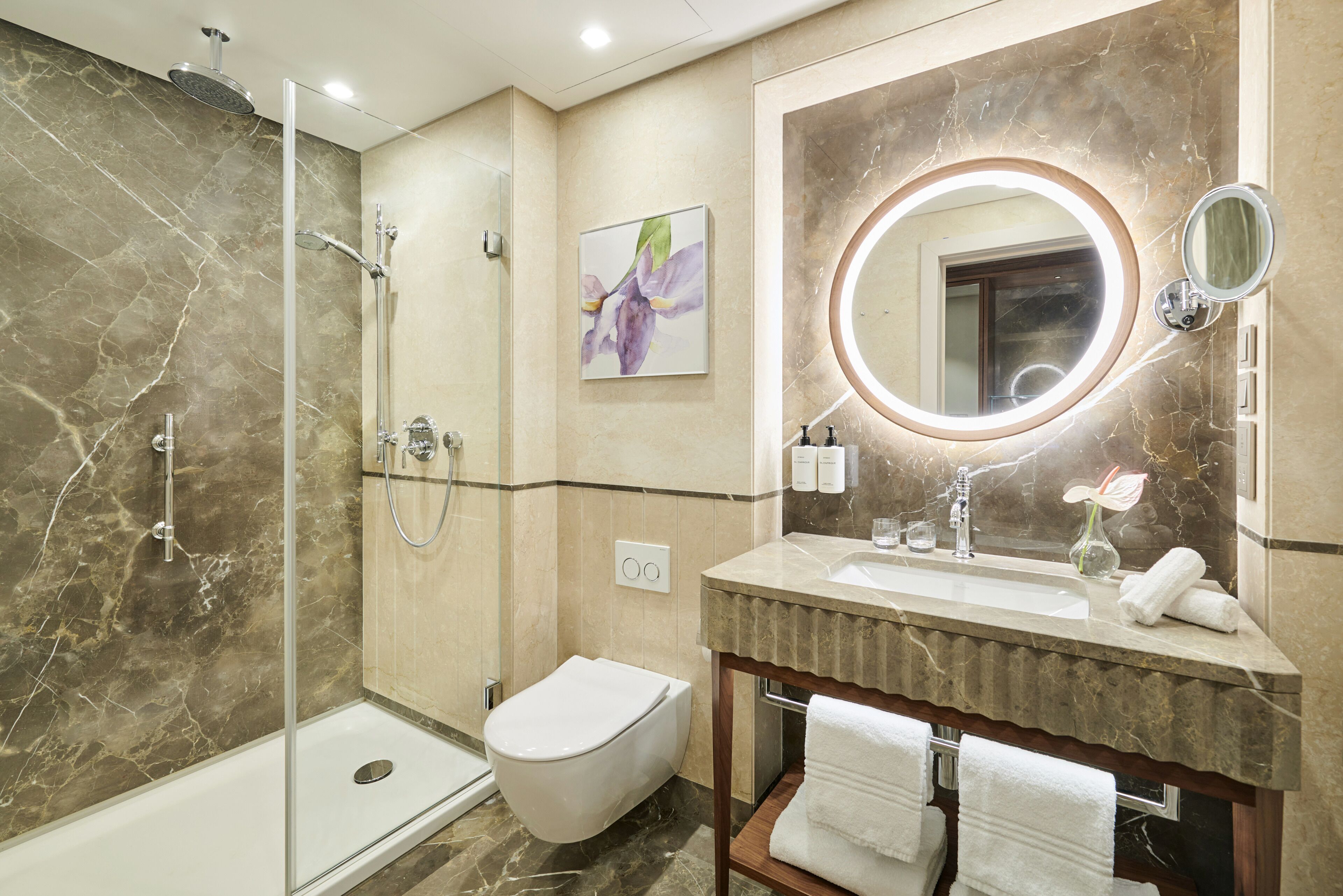 A modern bathroom with a glass-enclosed shower, a toilet, a sink with a marble countertop, and a large mirror with a circular frame.