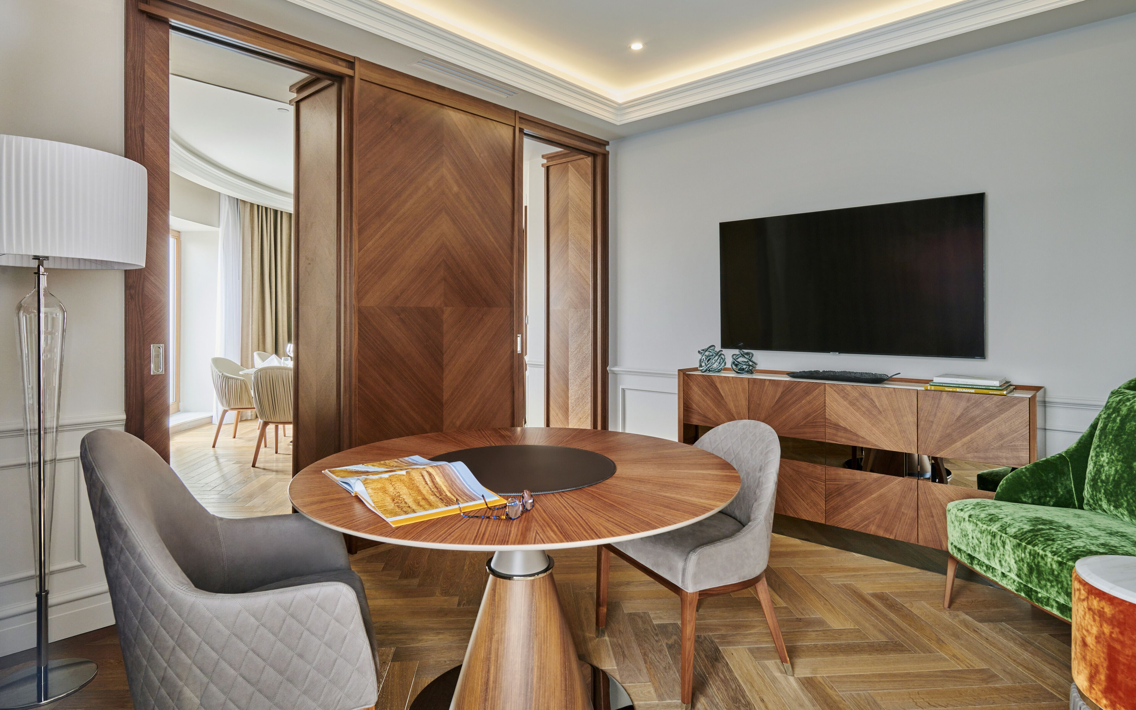 A modern room with a round wooden table, two grey chairs, a green sofa, a TV, and a wooden cabinet.