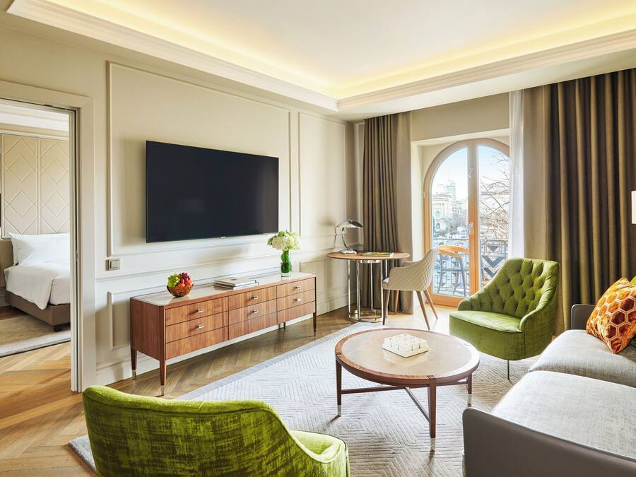 Habitación de hotel moderna con un televisor de pantalla plana grande, un sofá, dos sillones, una pequeña mesa redonda, una consola de madera con frutero, y vistas de la ciudad a través de un amplio ventanal.
