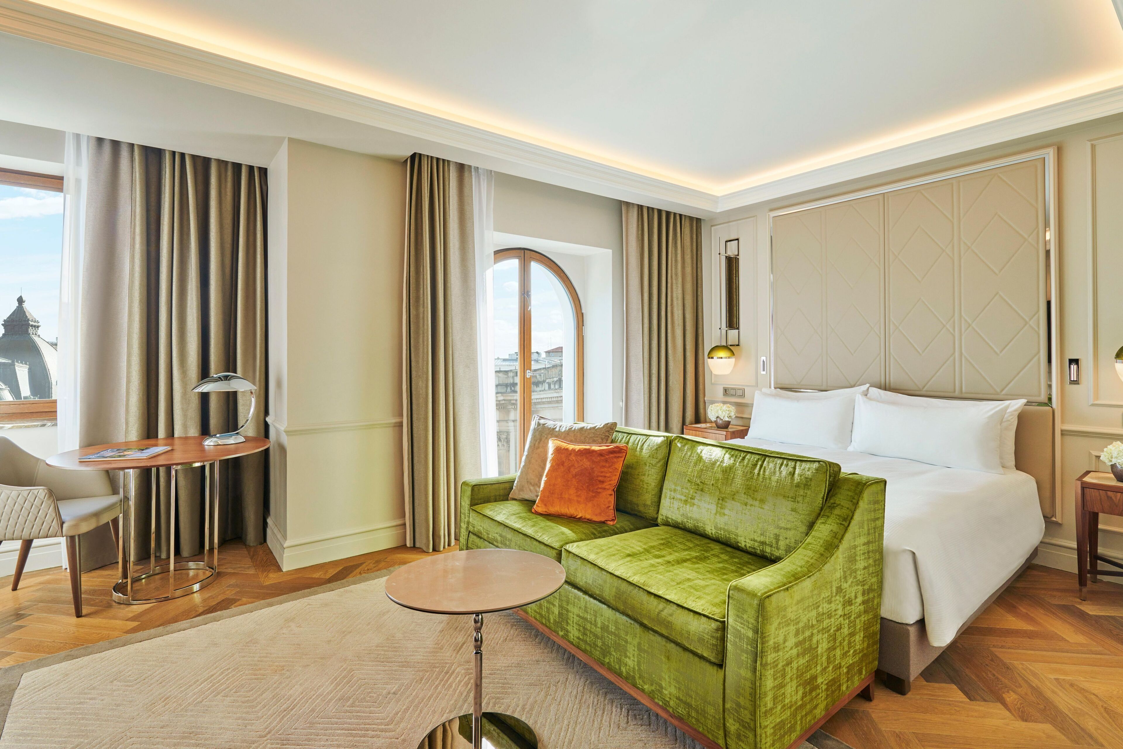 A hotel room with a green sofa, a round table, a bed, and a view of a cityscape through a window.