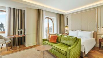 Camera d'albergo con un divano verde, un tavolo rotondo, un letto e vista sulla città attraverso una finestra.