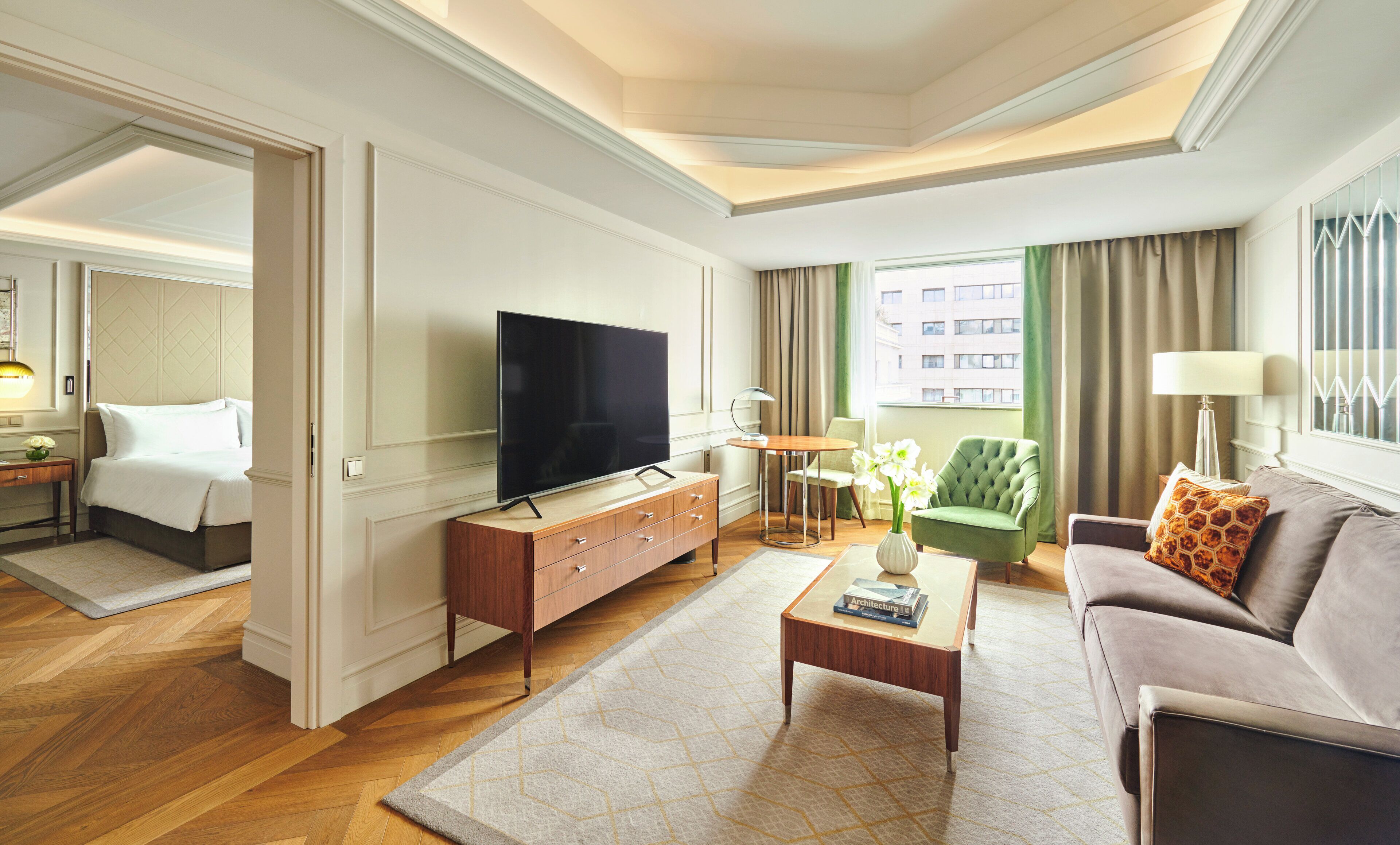 Ein Hotelzimmer mit Flachbildfernseher, einem Holztischchen, einer Couch, einem grünen Sessel und einem Fenster mit Vorhängen.