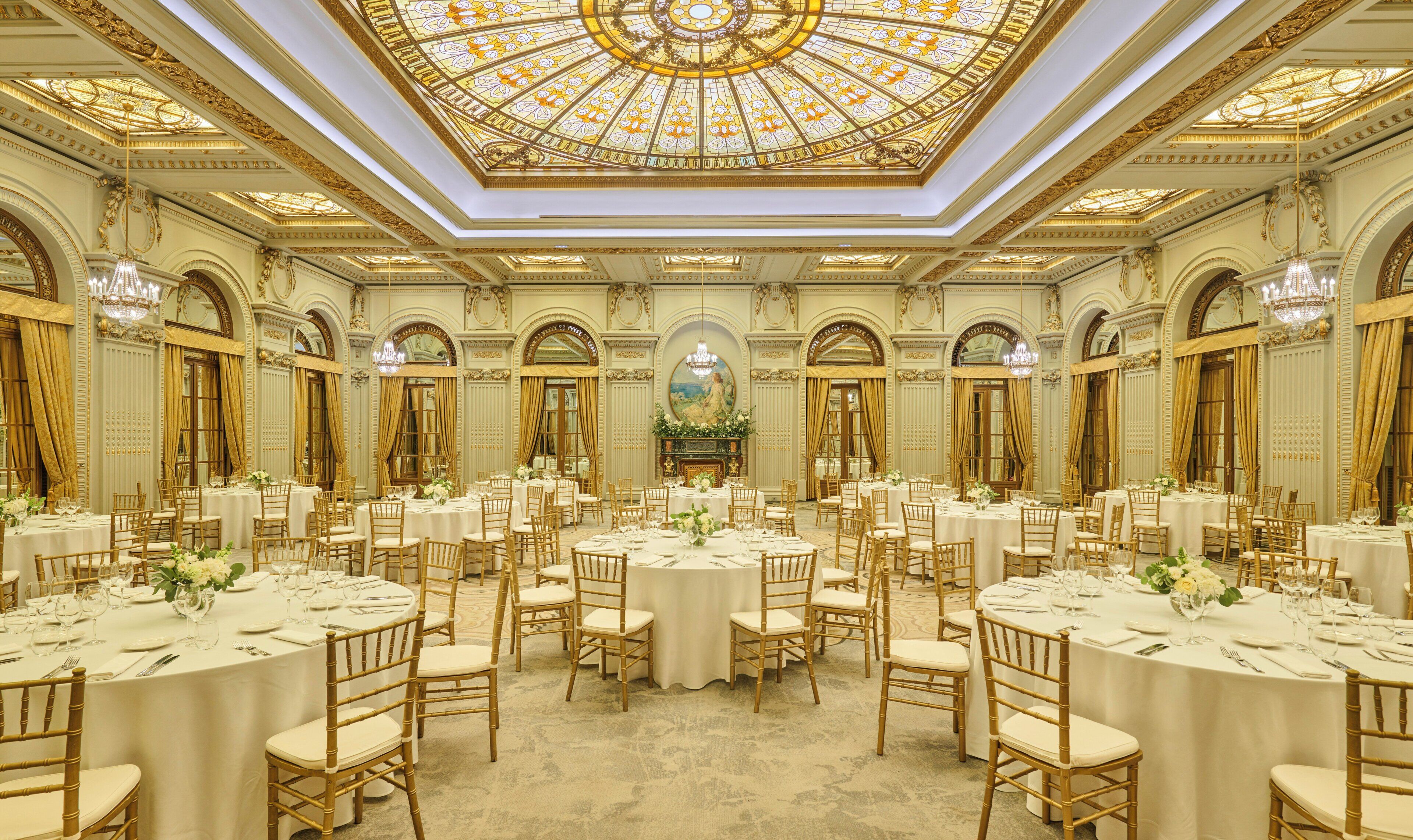 A grand banquet hall with multiple round tables, each set with white tablecloths and gold chairs.