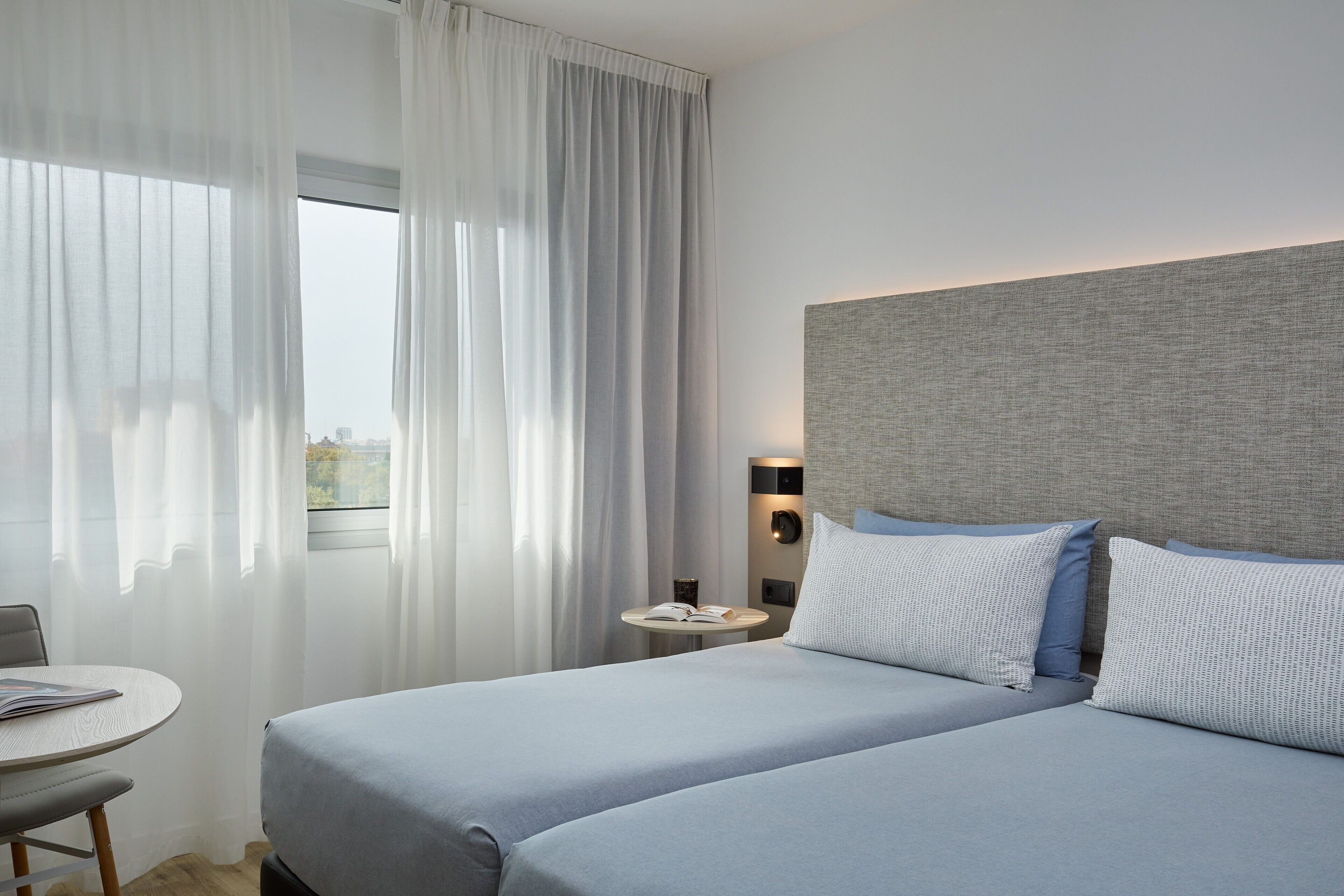 Ein Hotelzimmer mit einem Bett, einem kleinen Tisch mit einem Buch und einem Fenster mit Vorhängen.