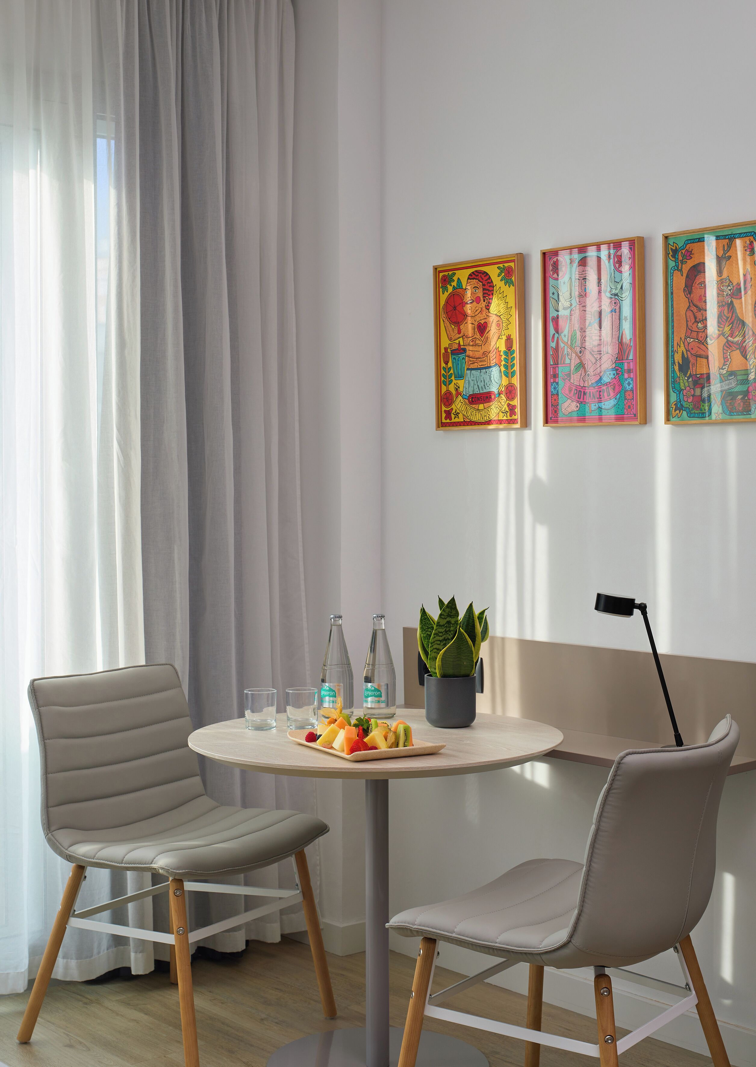 A modern room with a round table, two chairs, a potted plant, and three colorful artworks on the wall.